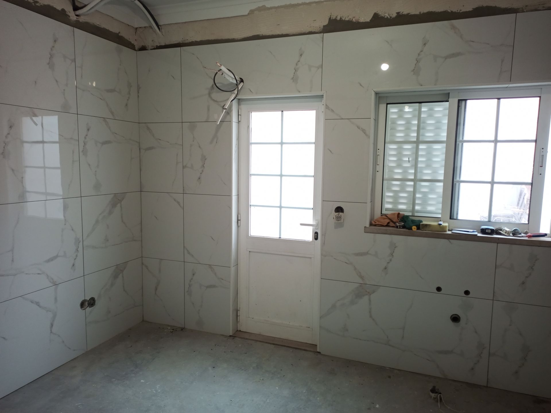 Interior de um cômodo em construção com azulejos brancos que imitam mármore, uma porta branca e uma janela.