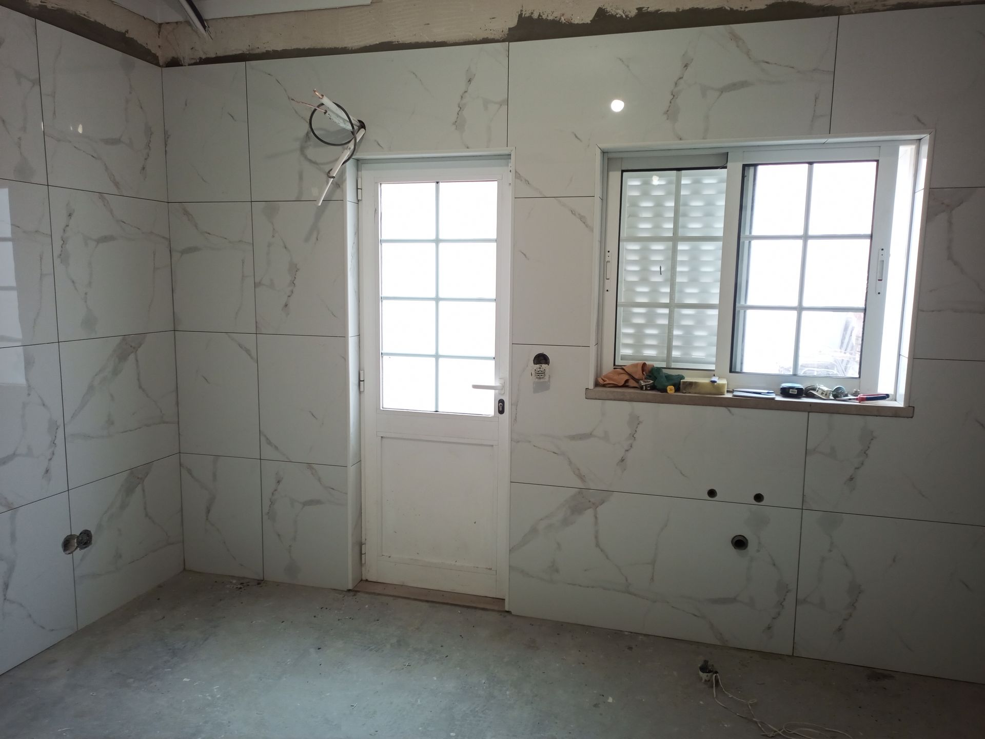 Quarto com azulejos brancos imitando mármore nas paredes, uma porta e uma janela com persianas; construção inacabada.