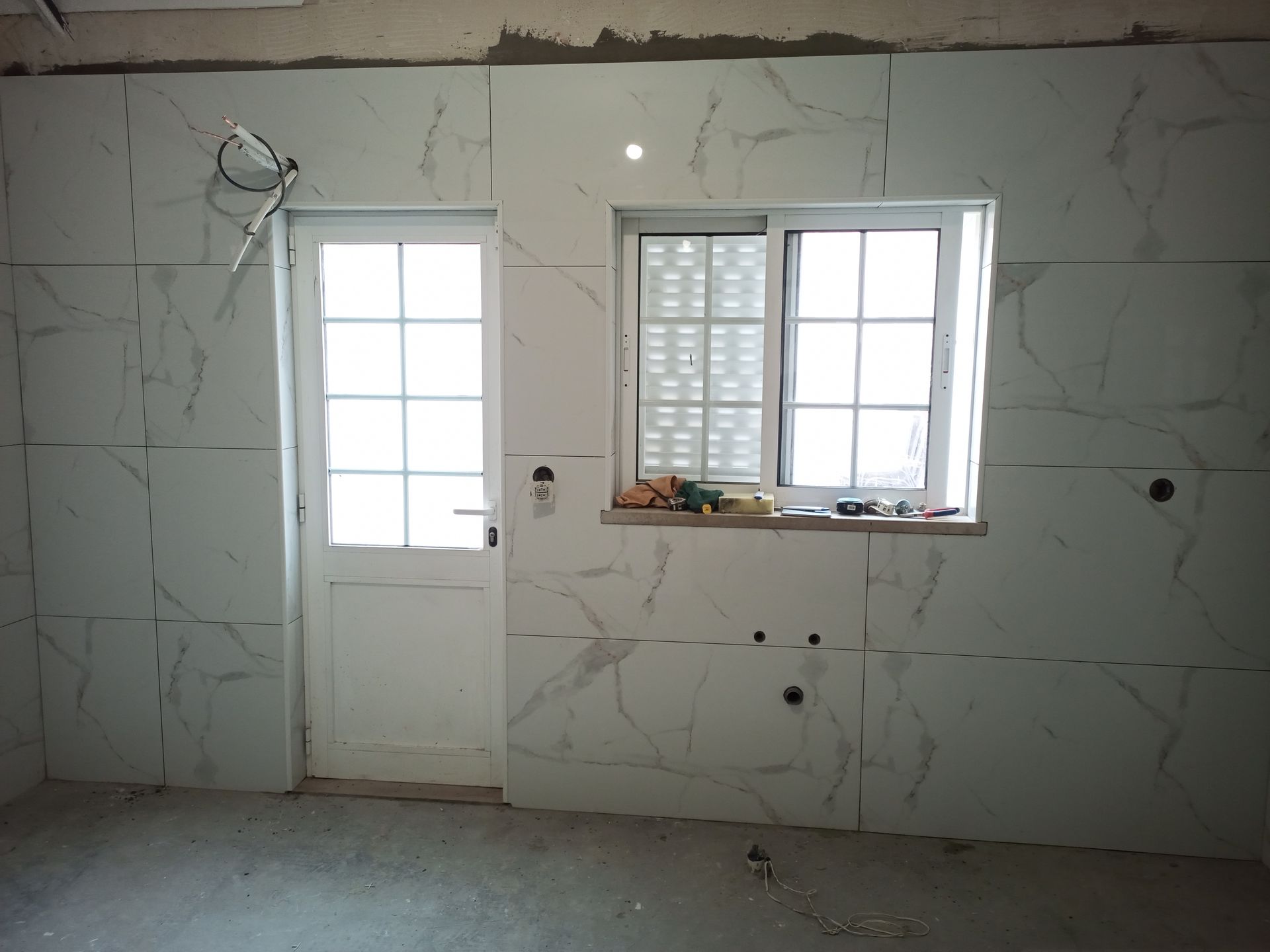 Quarto interior com paredes revestidas com azulejos brancos que imitam mármore, uma porta e uma janela; construção em andamento.