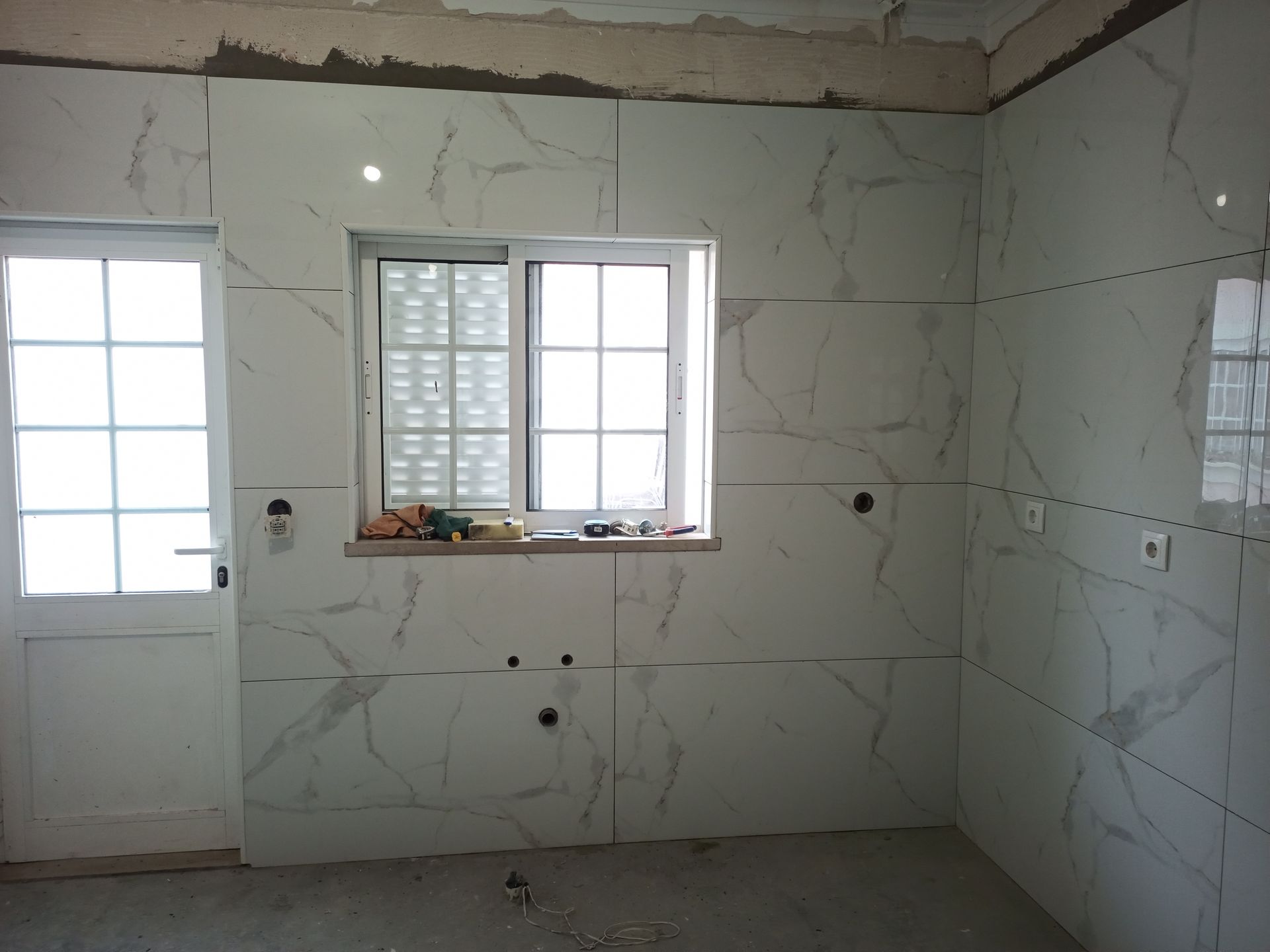 Quarto com paredes revestidas de azulejos brancos, janela e porta. O interior está em construção.