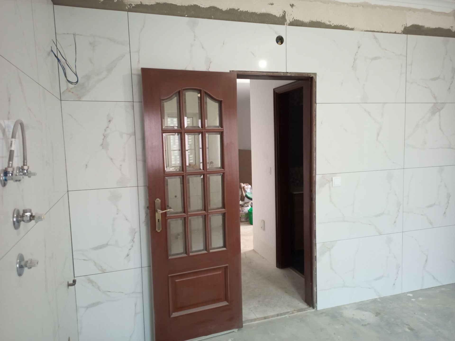 Porta marrom com painéis de vidro dá acesso a um cômodo com parede revestida de mármore branco e teto inacabado.