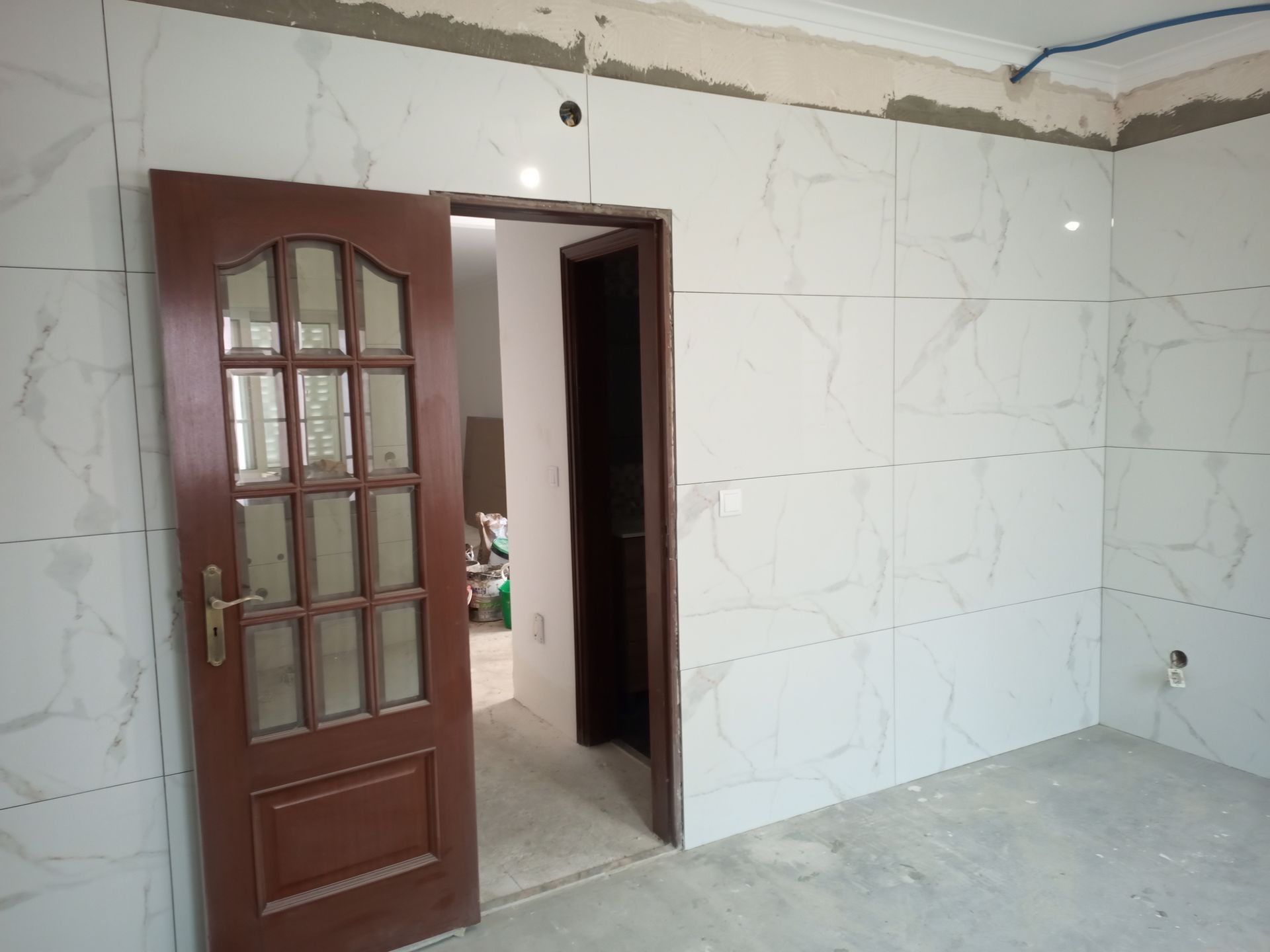 Quarto com paredes revestidas com azulejos brancos que imitam mármore, uma porta marrom com painéis de vidro e uma porta aberta para outro cômodo.