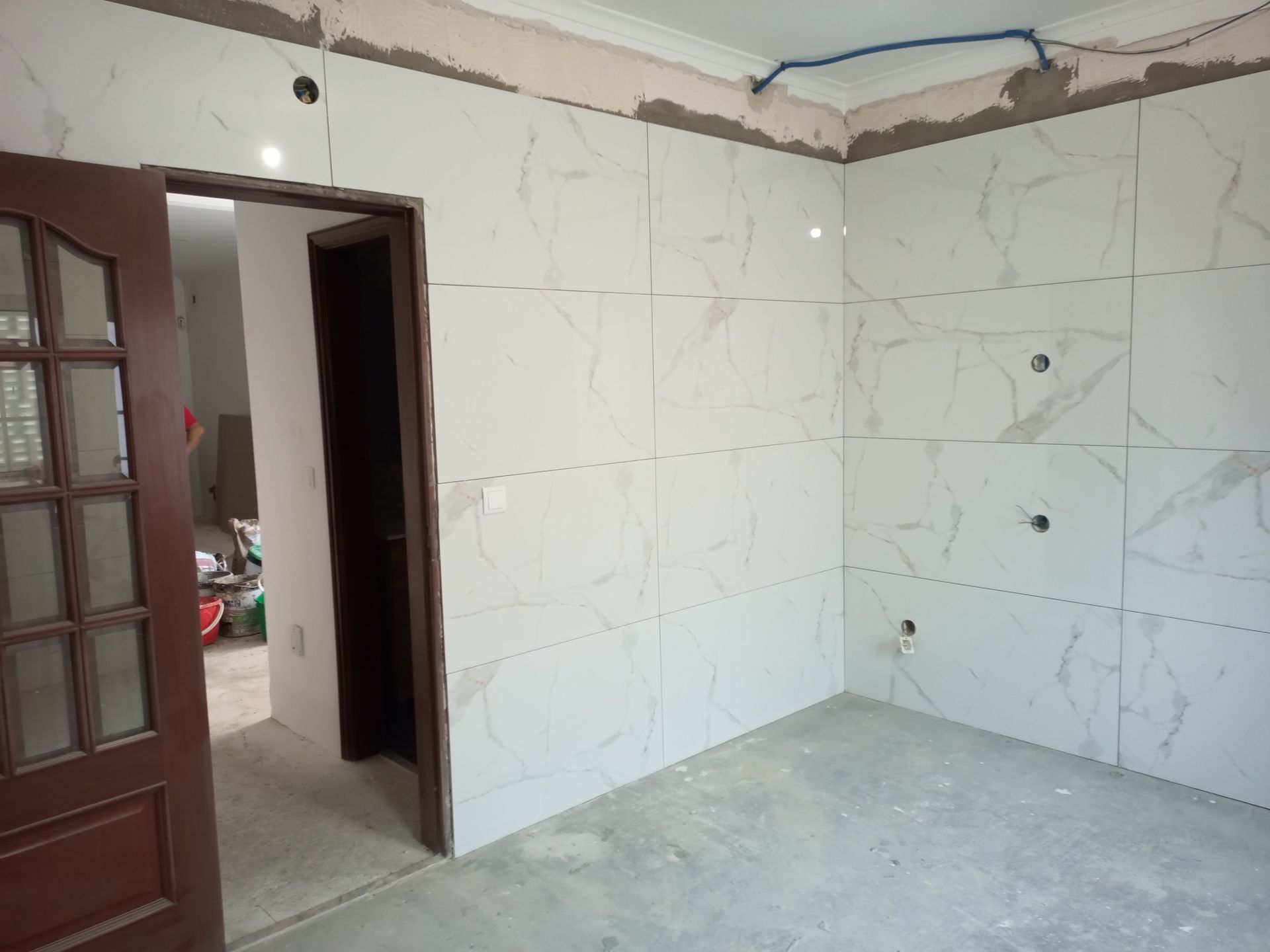 Quarto com piso recém-azulejado e portas abertas. Azulejos brancos, porta marrom, piso inacabado, cano azul à mostra.