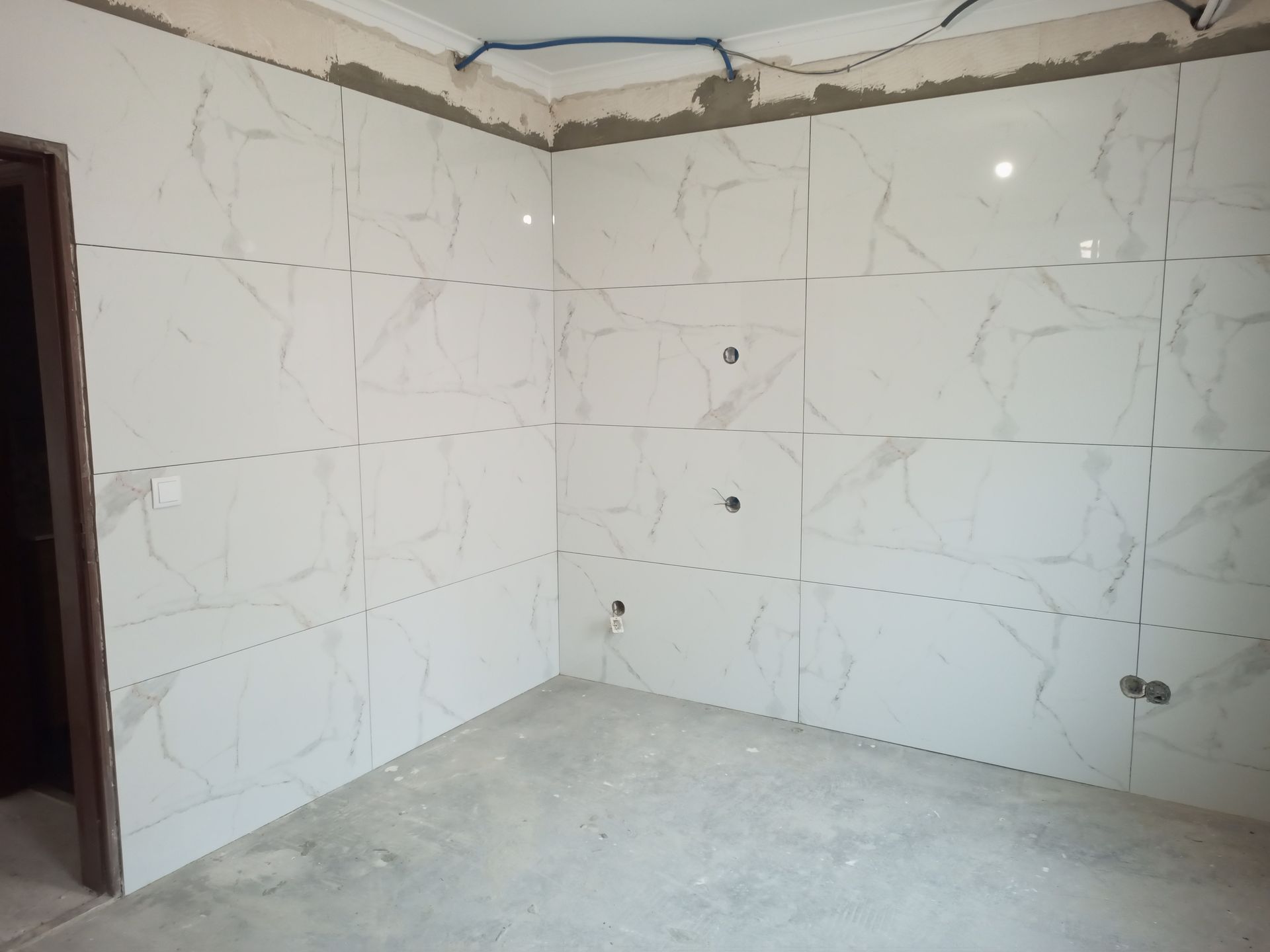 Paredes revestidas com azulejos brancos de um cômodo em construção, com fiação aparente e piso inacabado.