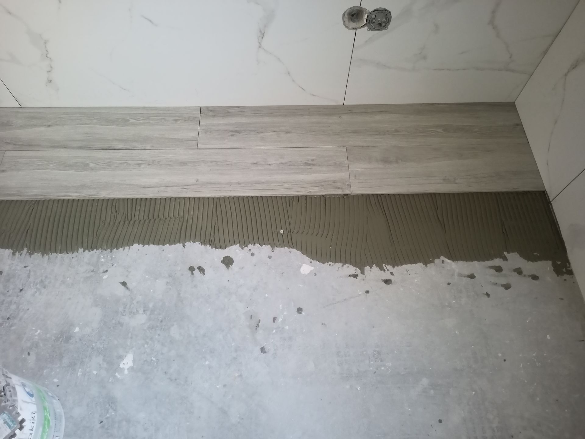 Instalação de revestimento cerâmico imitando madeira em tons claros na parede, sobre a camada de argamassa e o piso de concreto inacabado.