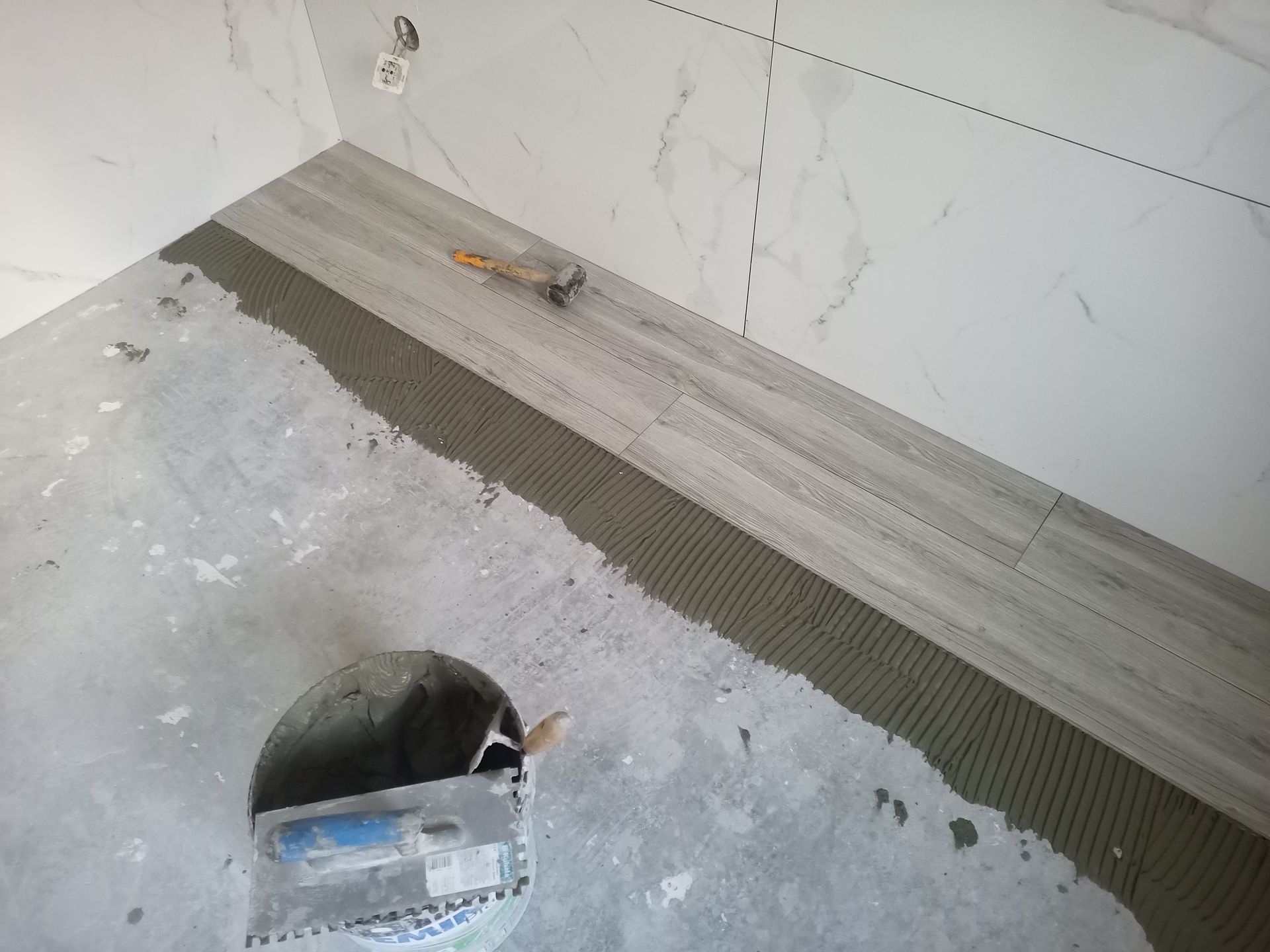 Assentamento de piso de banheiro com azulejos que imitam madeira; balde de argamassa e ferramentas visíveis.