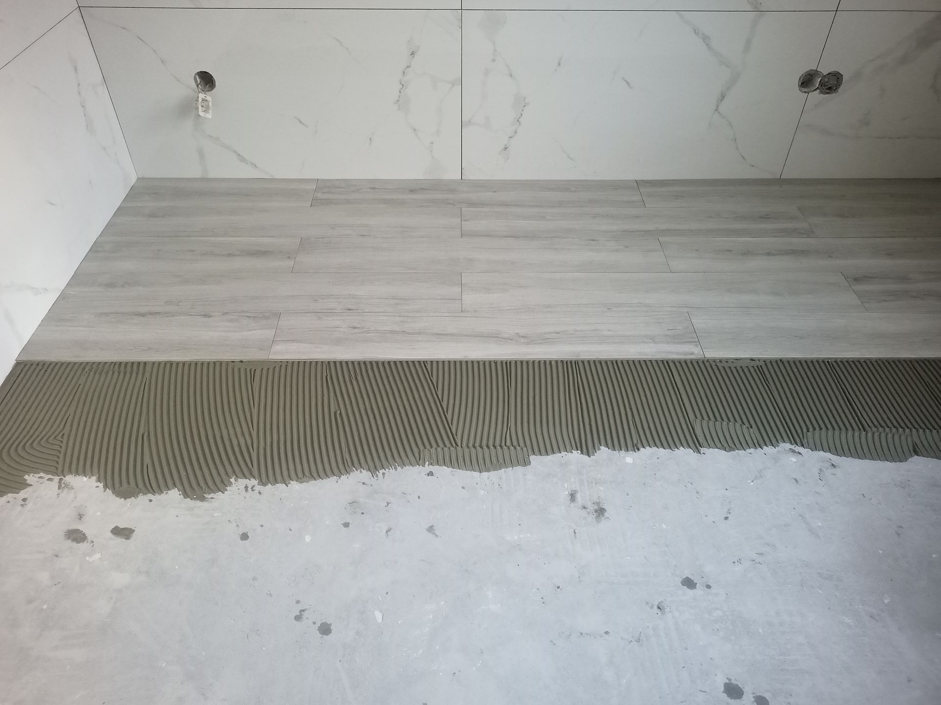 Instalação de azulejos no piso do banheiro em andamento; azulejos cinza imitando madeira assentados sobre adesivo, paredes e teto brancos.