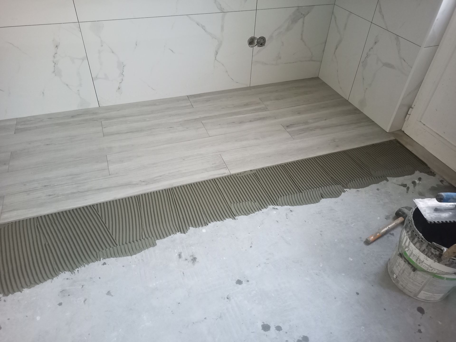 Piso do banheiro em processo de instalação de azulejos. Argamassa cinza sendo aplicada sobre o piso que imita madeira. Azulejos brancos que imitam mármore nas paredes.