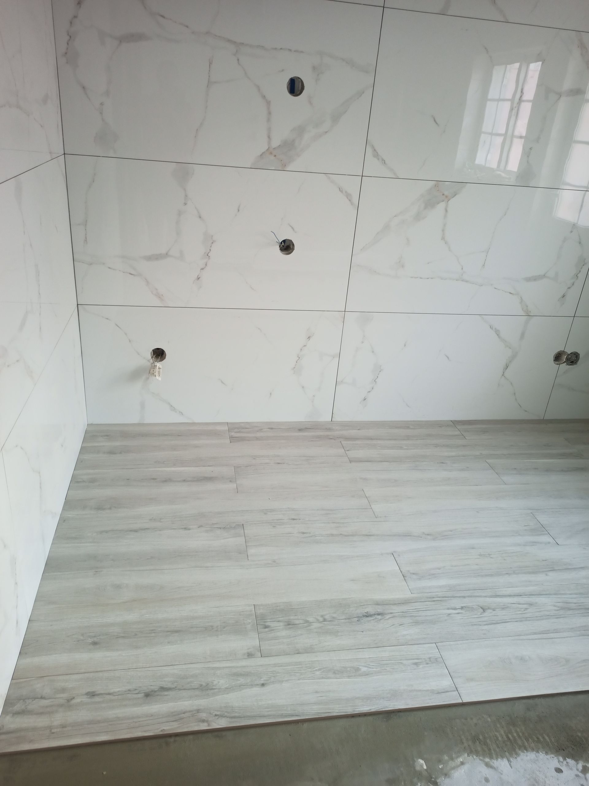 Interior de banheiro com paredes revestidas com azulejos brancos imitando mármore e piso com aparência de madeira em tom cinza claro.