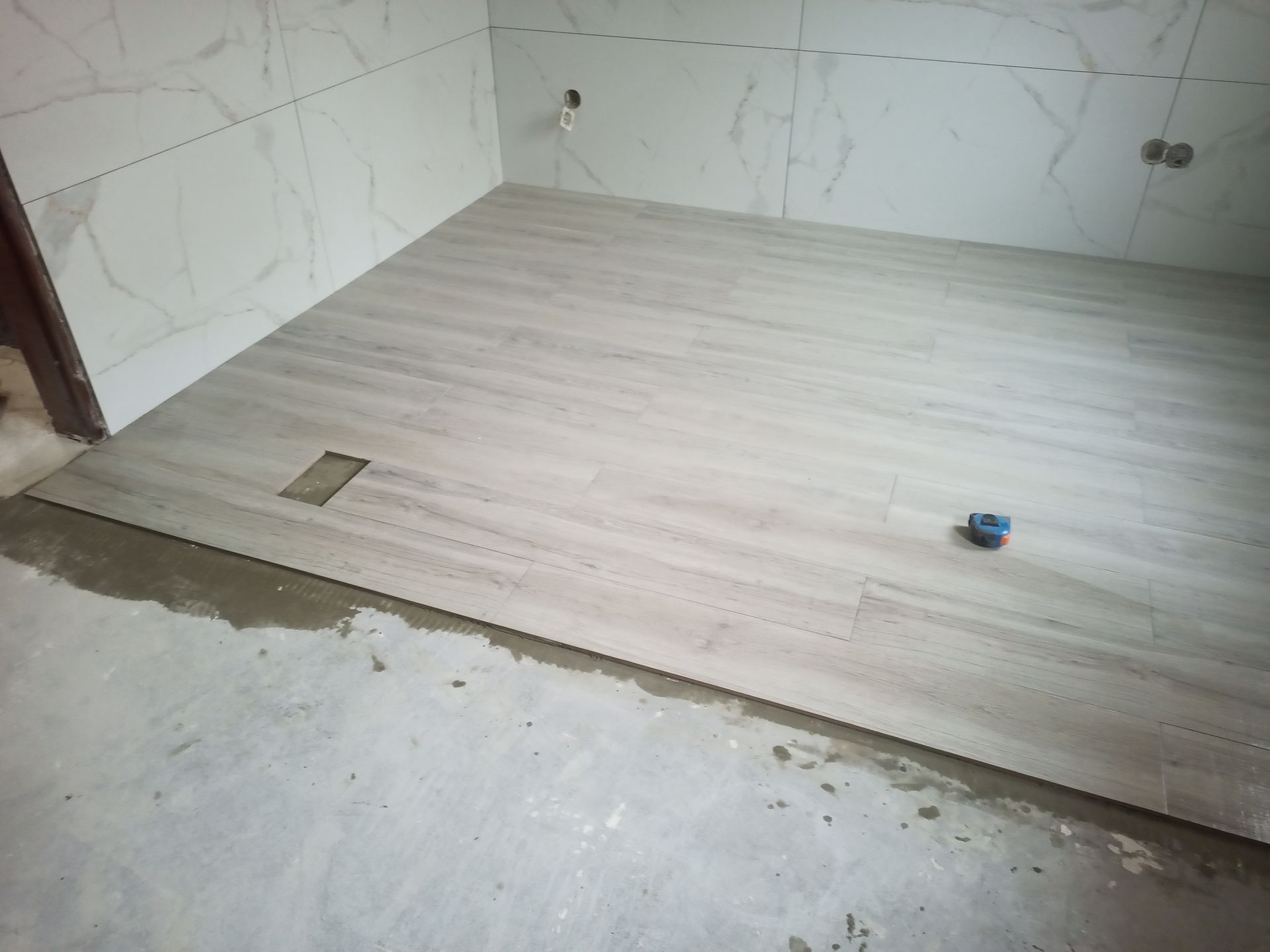 Instalação de azulejos no piso do banheiro em andamento; piso cerâmico com aparência de madeira clara já foi aplicado, e as paredes estão parcialmente revestidas com mármore.