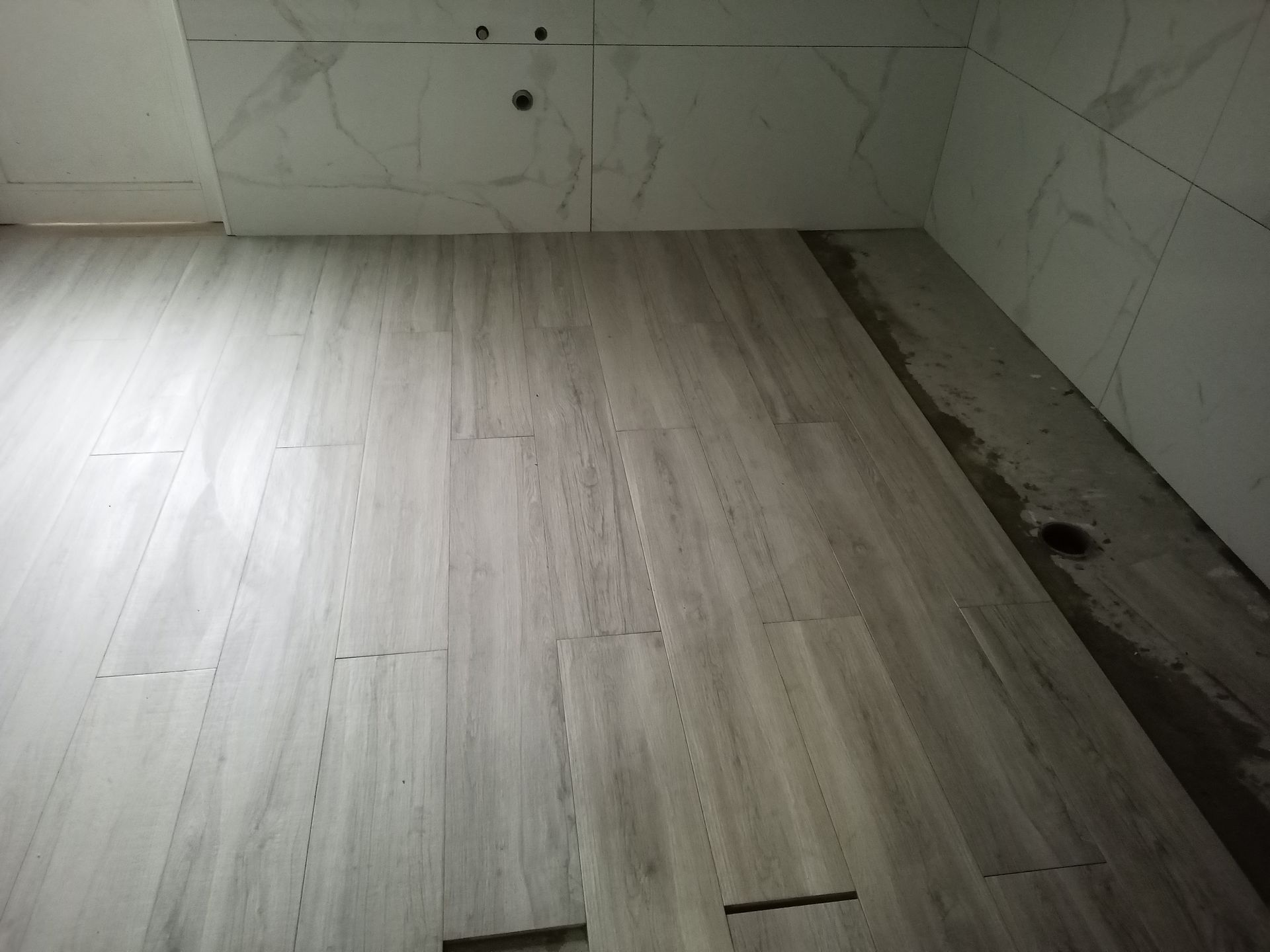 Banheiro com piso imitando madeira clara e paredes revestidas com azulejos brancos; construção parcialmente concluída.