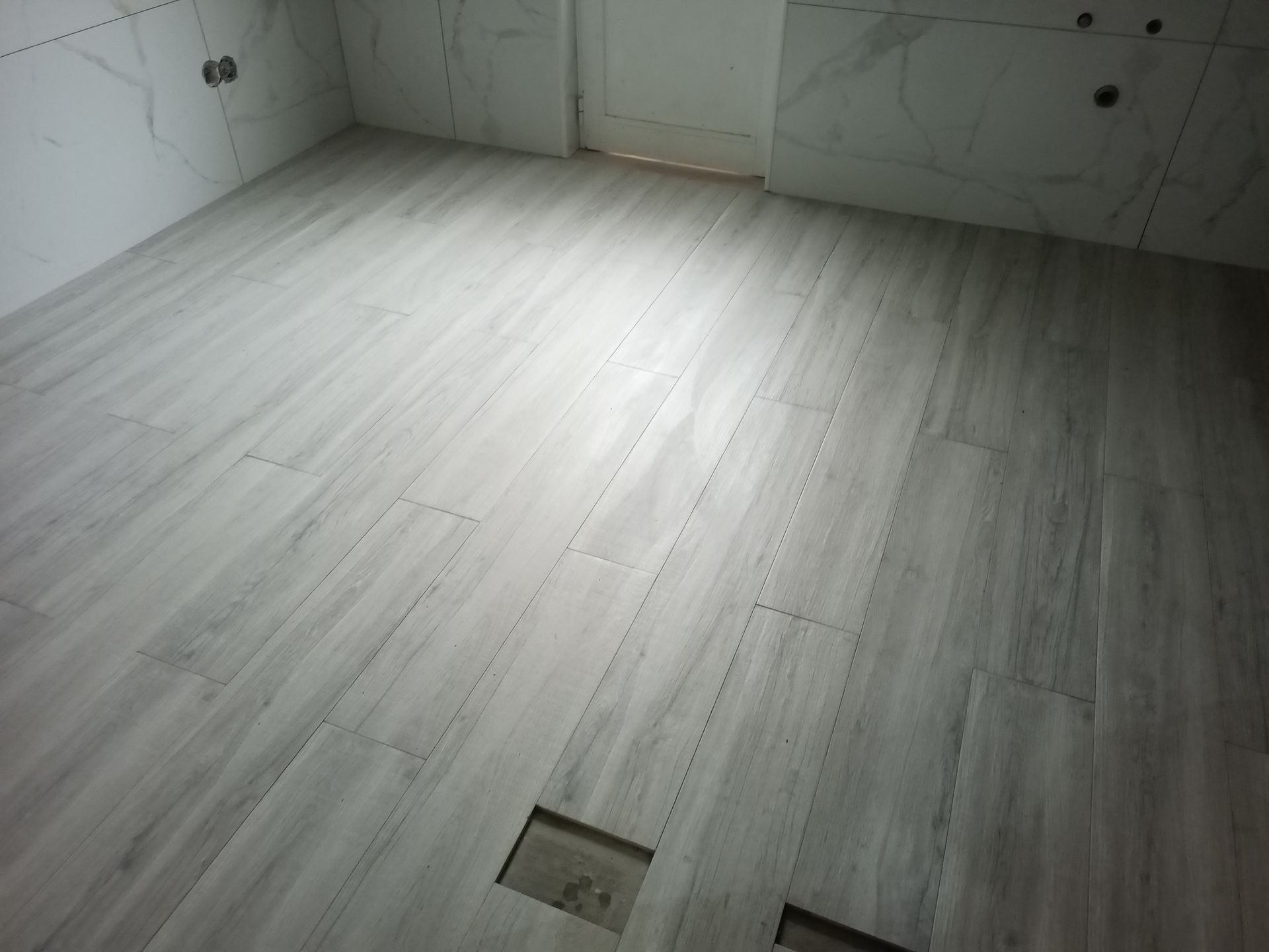 Piso de cerâmica imitando madeira cinza em um cômodo, com azulejos de parede que imitam mármore. As peças retangulares de cerâmica são assentadas com juntas visíveis.