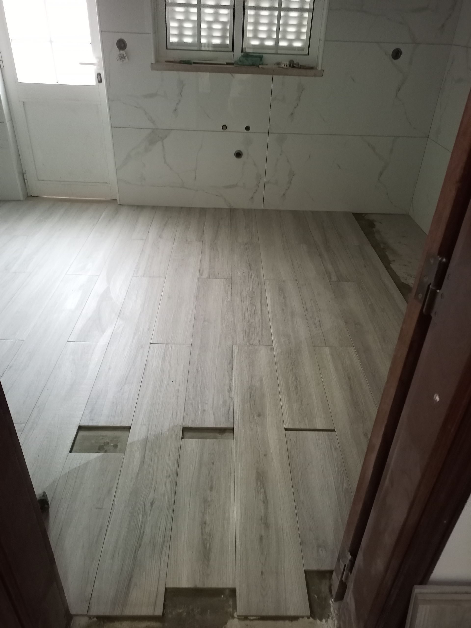 Quarto com piso de azulejo cinza claro e paredes revestidas com azulejos brancos; janela ao fundo. Faltam vários azulejos.