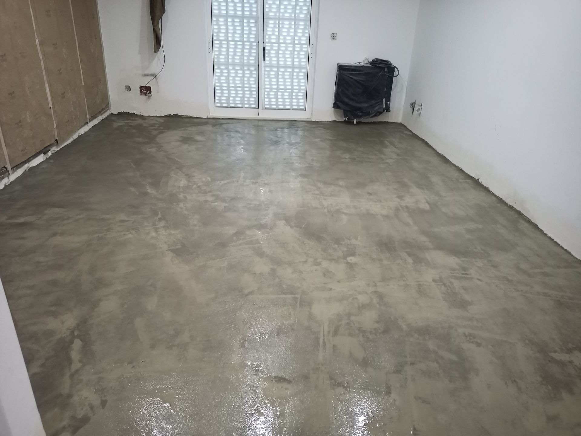 Quarto com piso de concreto cinza e molhado; paredes brancas; e porta de vidro fechada.