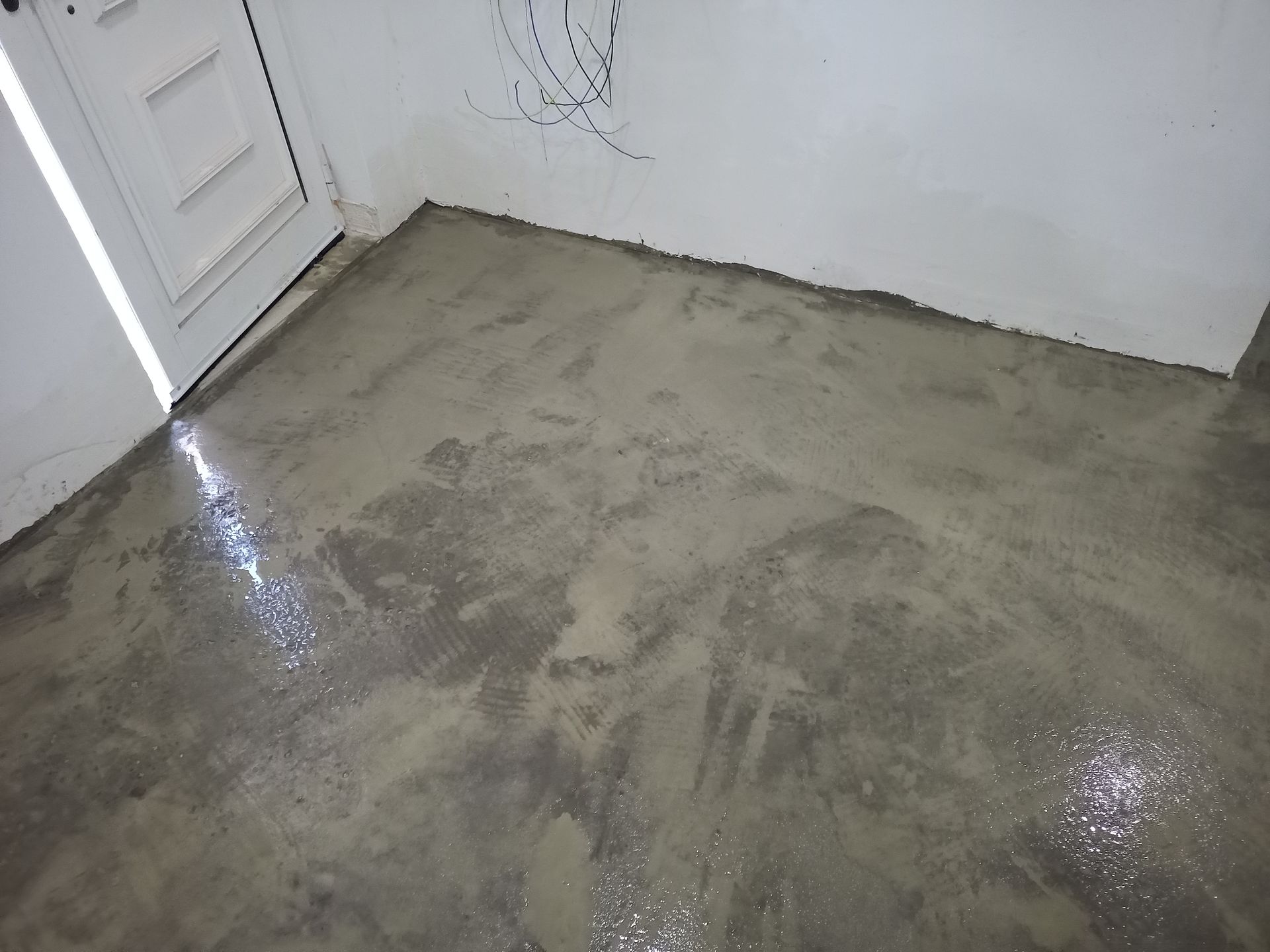Piso de concreto cinza com gotas de água, próximo a uma porta e parede brancas.