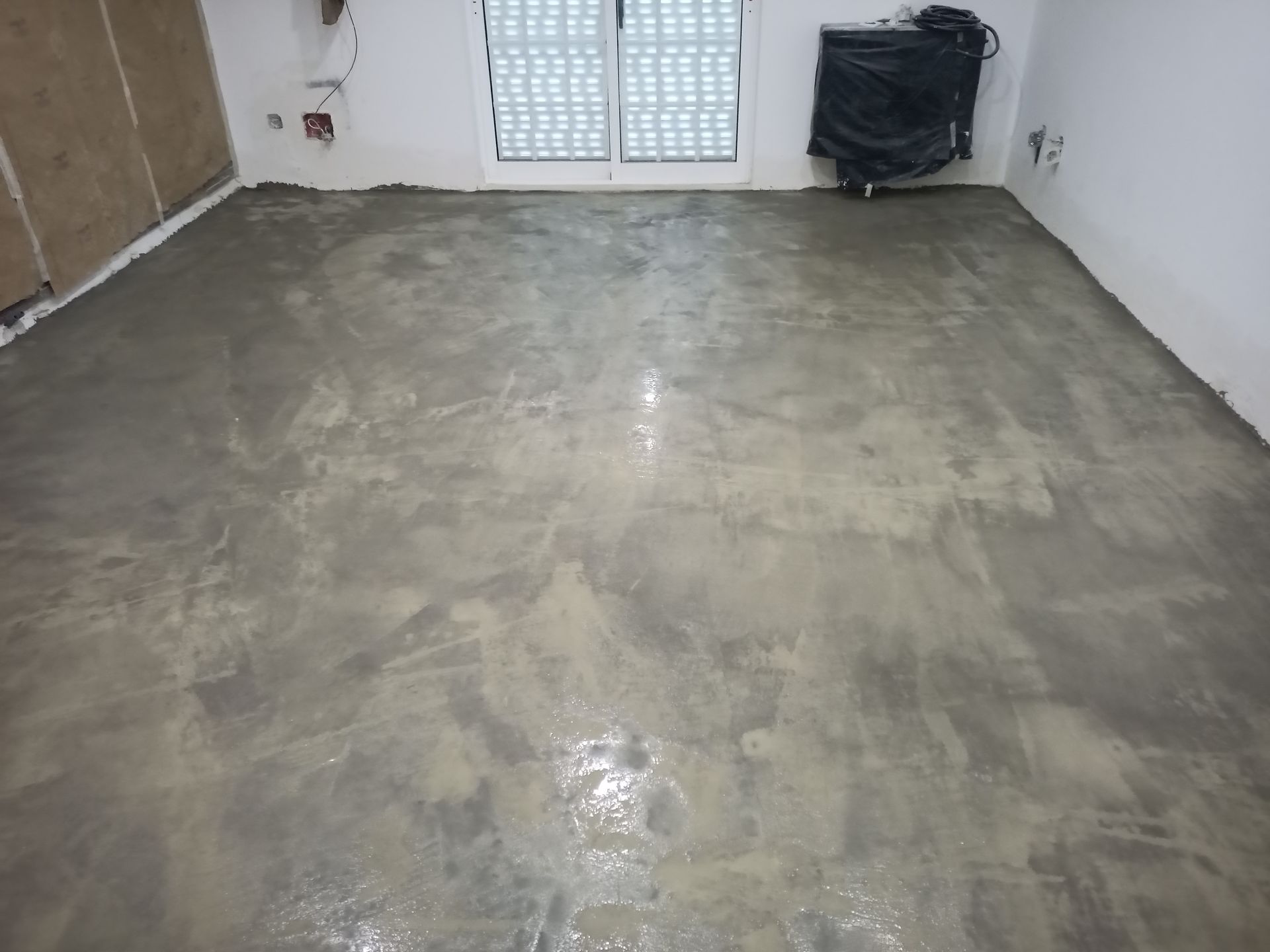 Quarto com piso de concreto cinza e molhado. As paredes são brancas, com uma janela e um objeto coberto.