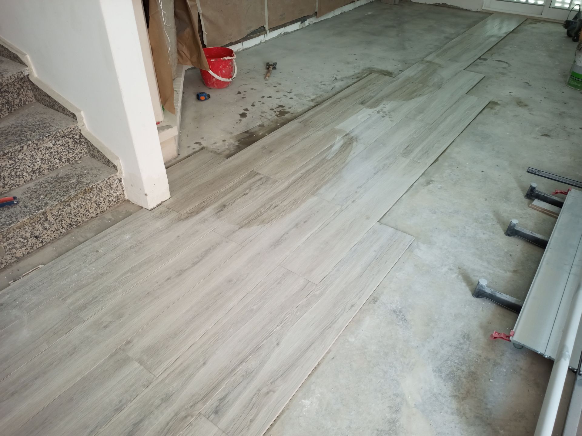 Colocação de piso em andamento; ladrilhos com aparência de madeira assentados sobre piso de concreto, próximo à escada, ferramentas e balde visíveis.
