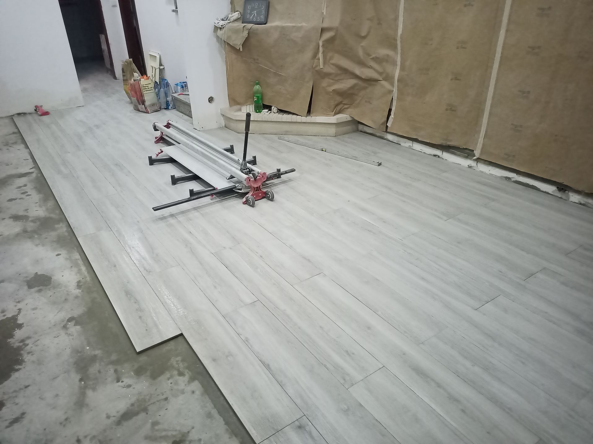 Assentamento de ladrilhos cinza imitando madeira. Uma máquina de cortar ladrilhos está sobre os ladrilhos recém-assentados em um cômodo em construção.