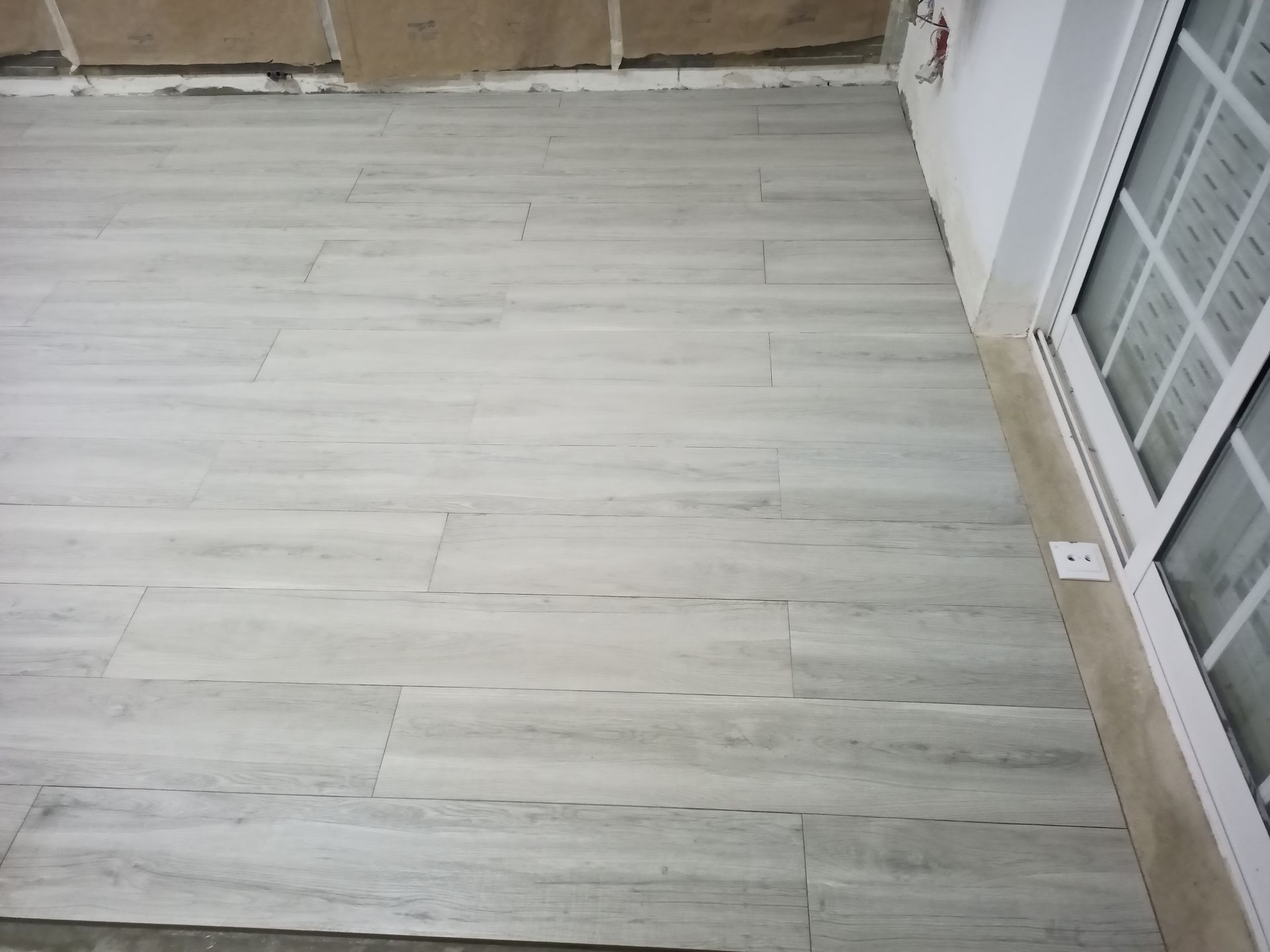 Piso de azulejo imitando madeira em tom cinza claro, instalado próximo a uma porta de vidro deslizante e a uma parede.