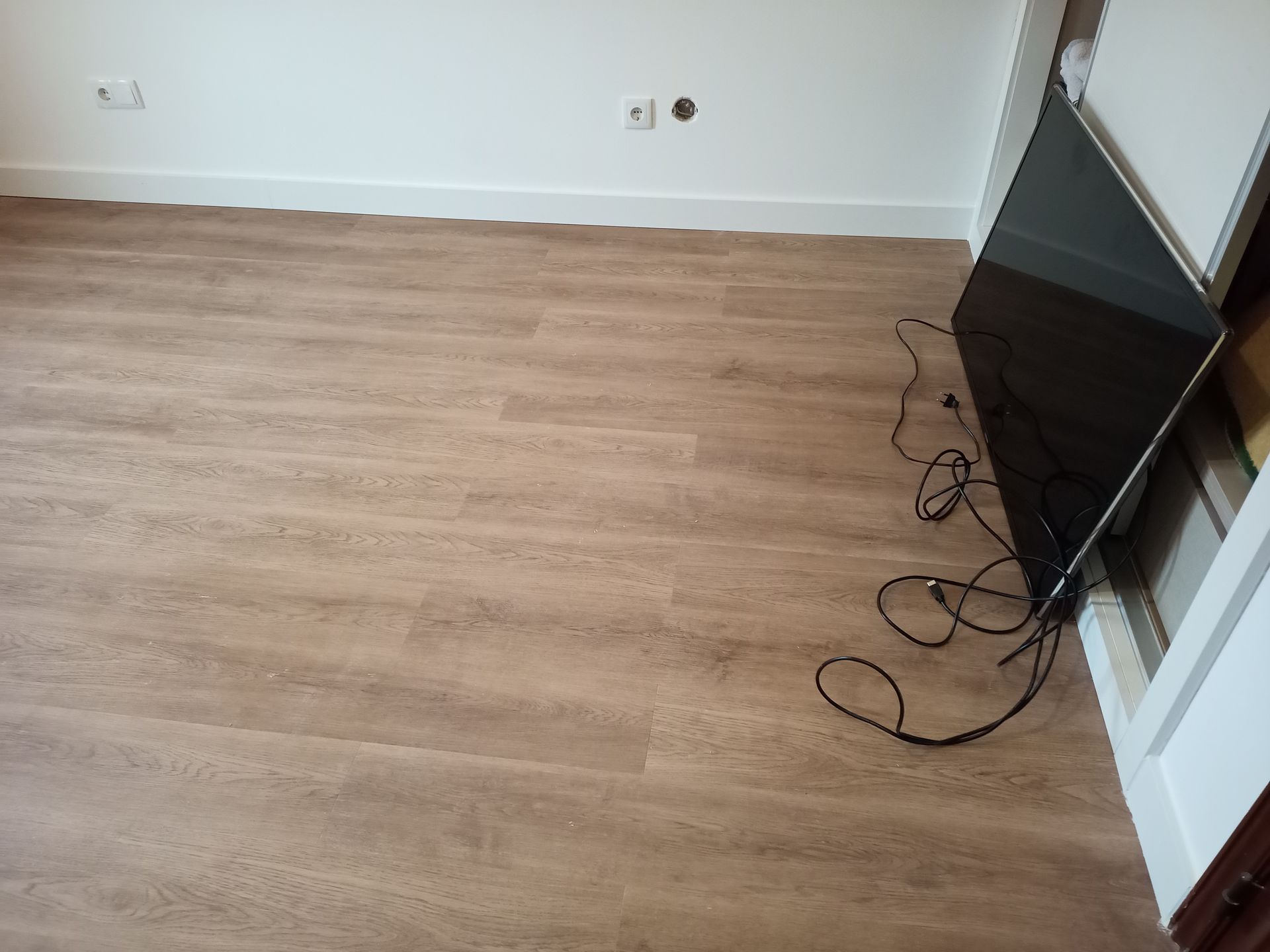 Quarto vazio com piso de madeira, molduras brancas e uma televisão encostada no batente da porta.