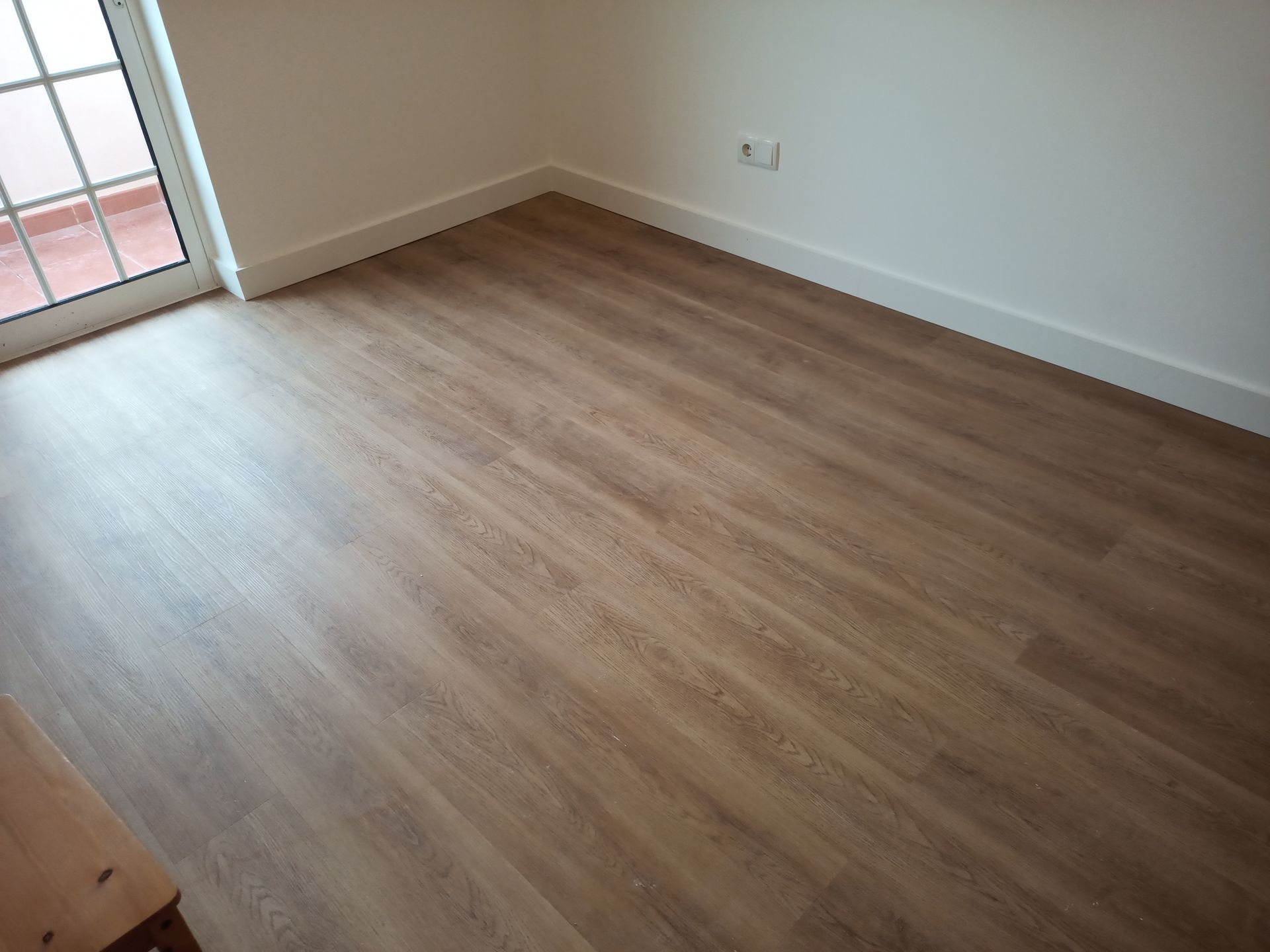 Quarto com piso imitando madeira em tom castanho claro e paredes brancas, com uma porta à esquerda.