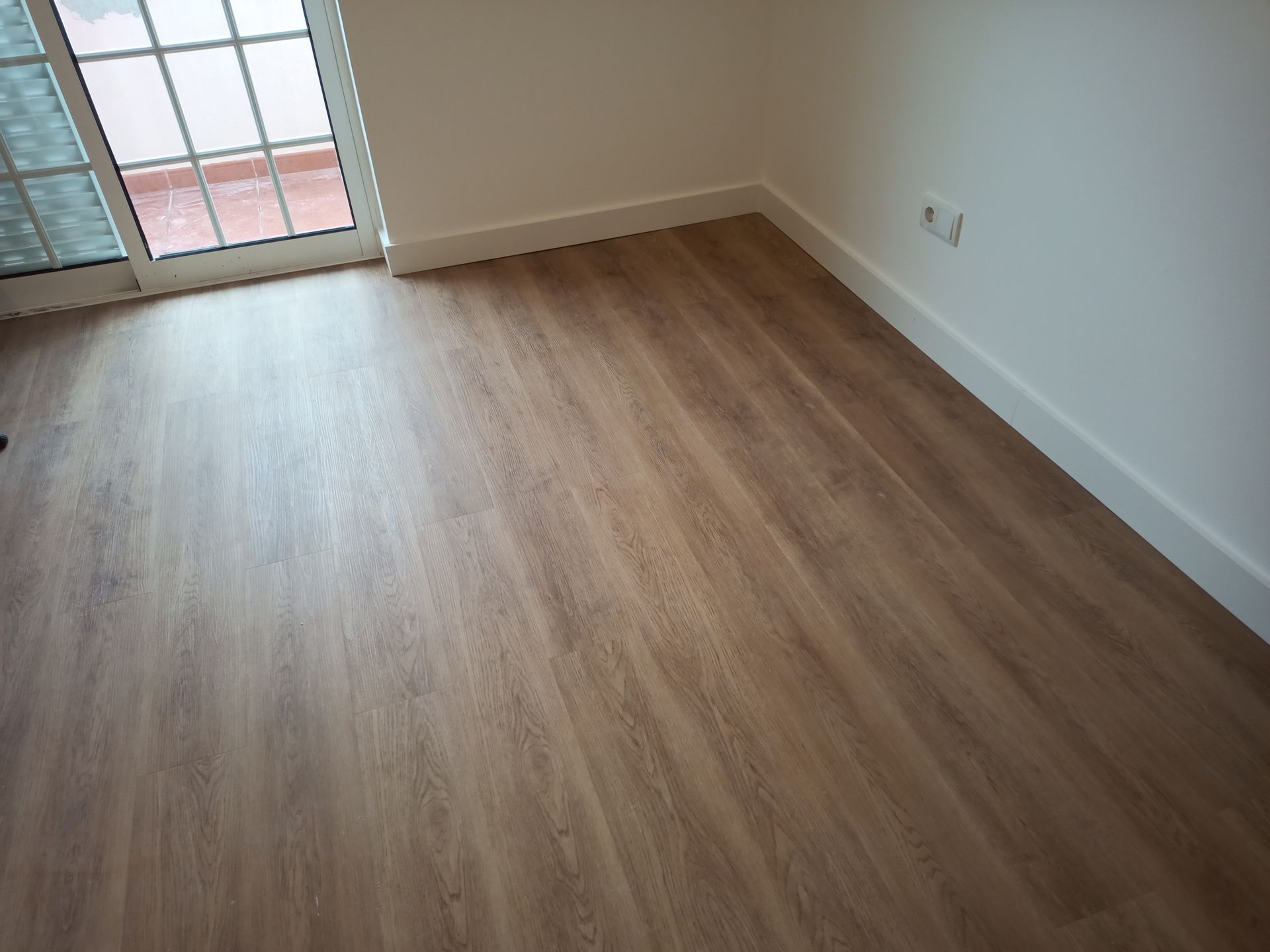 Quarto com piso de madeira, rodapés brancos e porta de vidro deslizante.