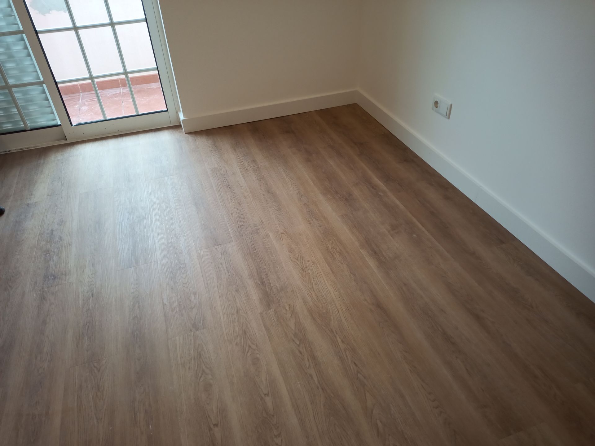 Quarto com piso imitando madeira em tom castanho claro, rodapés brancos e uma parede branca com uma tomada elétrica. Uma porta de vidro é visível.