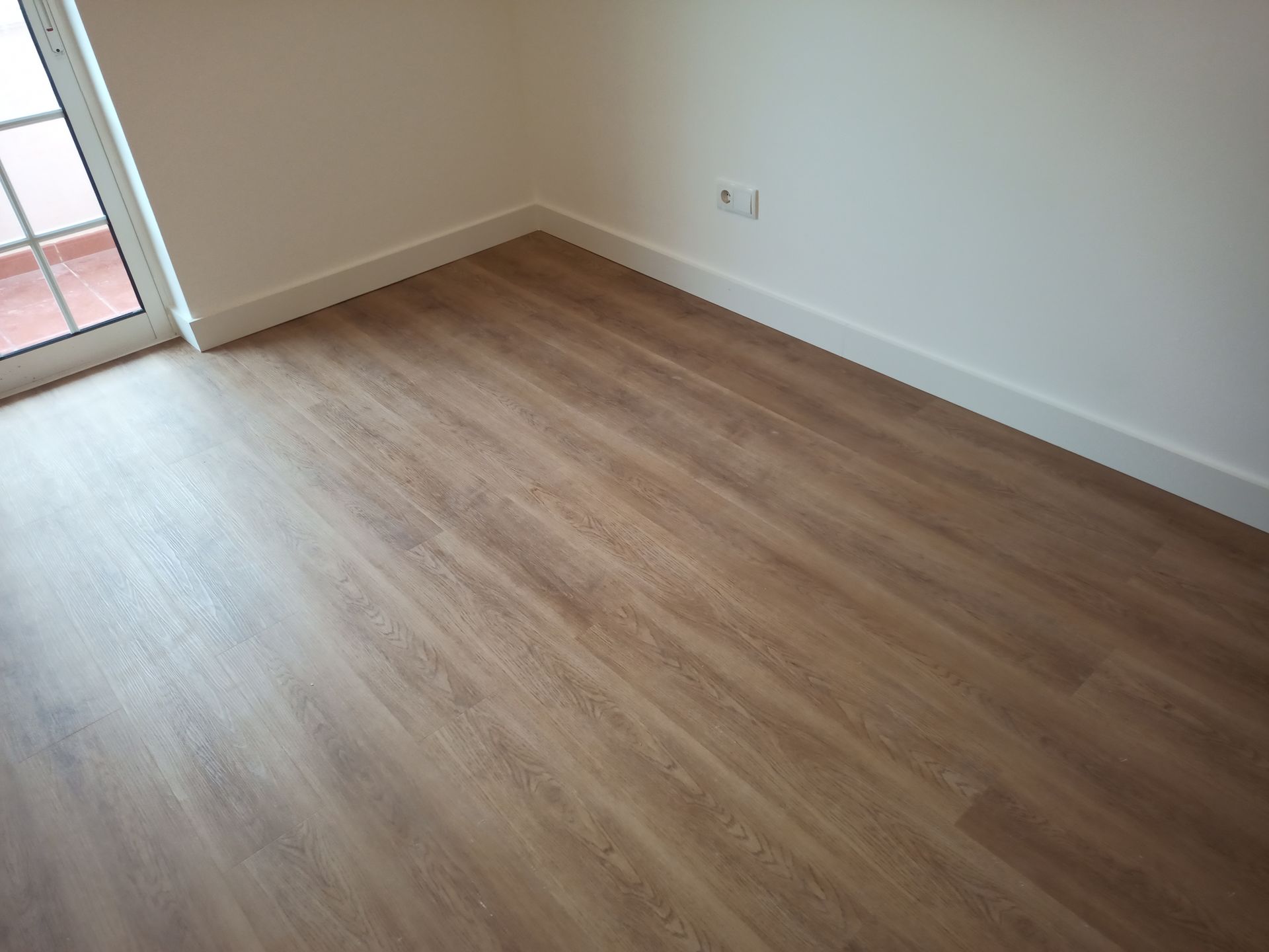 Piso de madeira em um quarto com paredes brancas, rodapés e uma porta que dá para uma varanda.