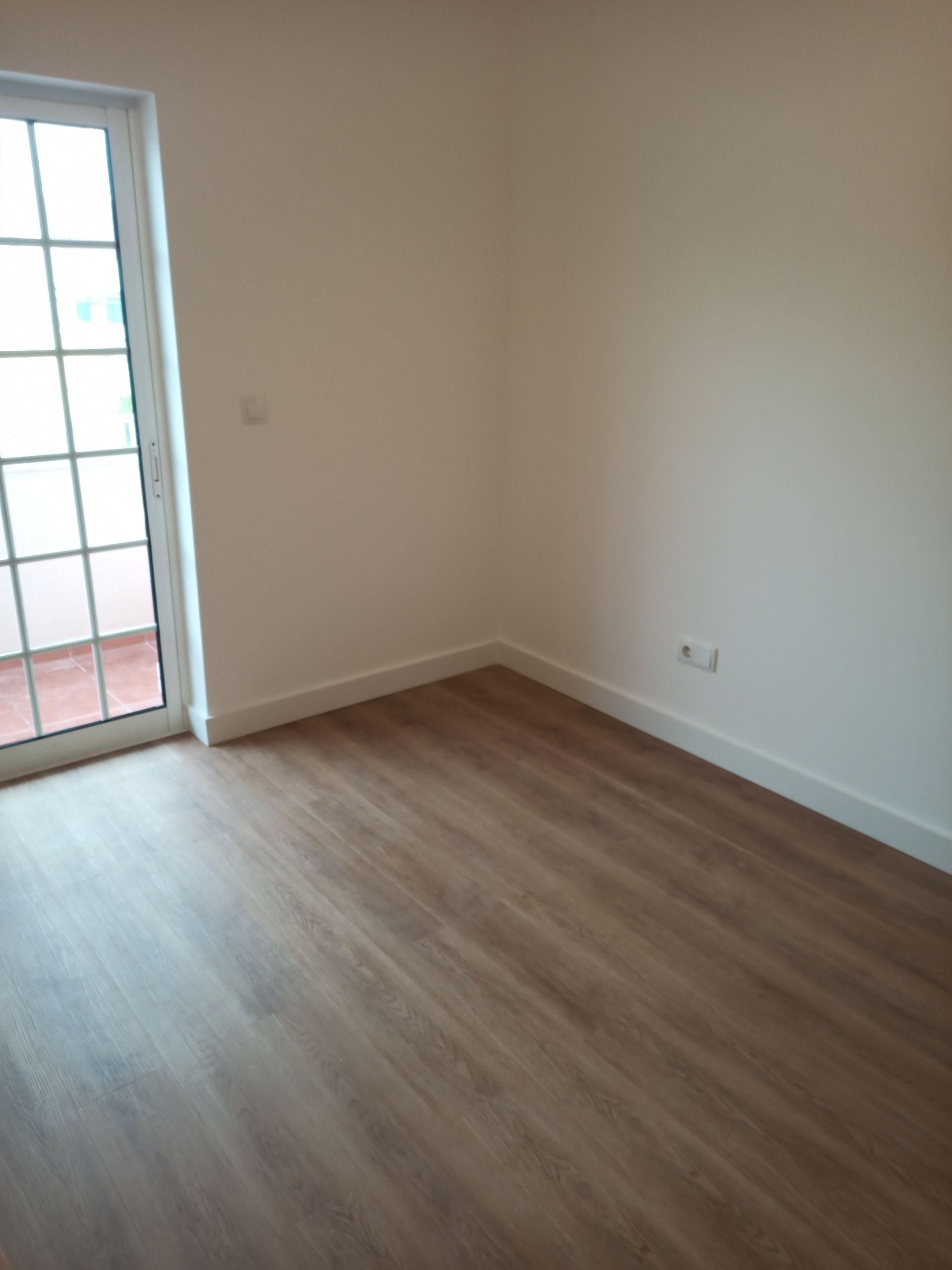 Quarto vazio com piso de madeira, paredes brancas e porta de vidro.