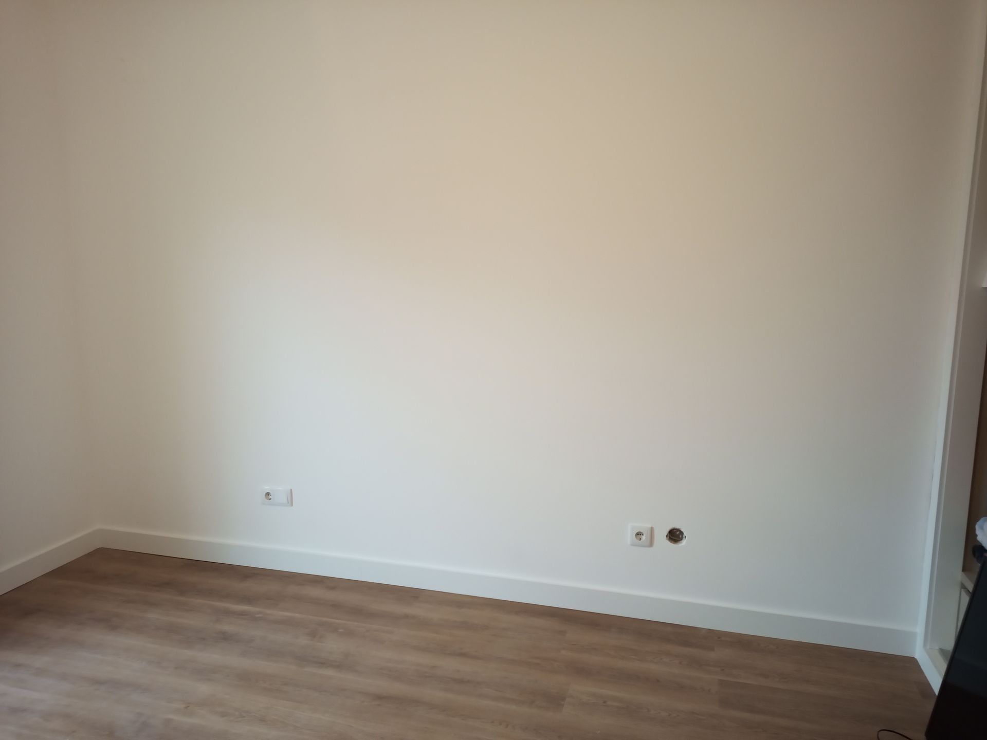 Quarto vazio com paredes brancas, piso de madeira e rodapés; tomadas visíveis.