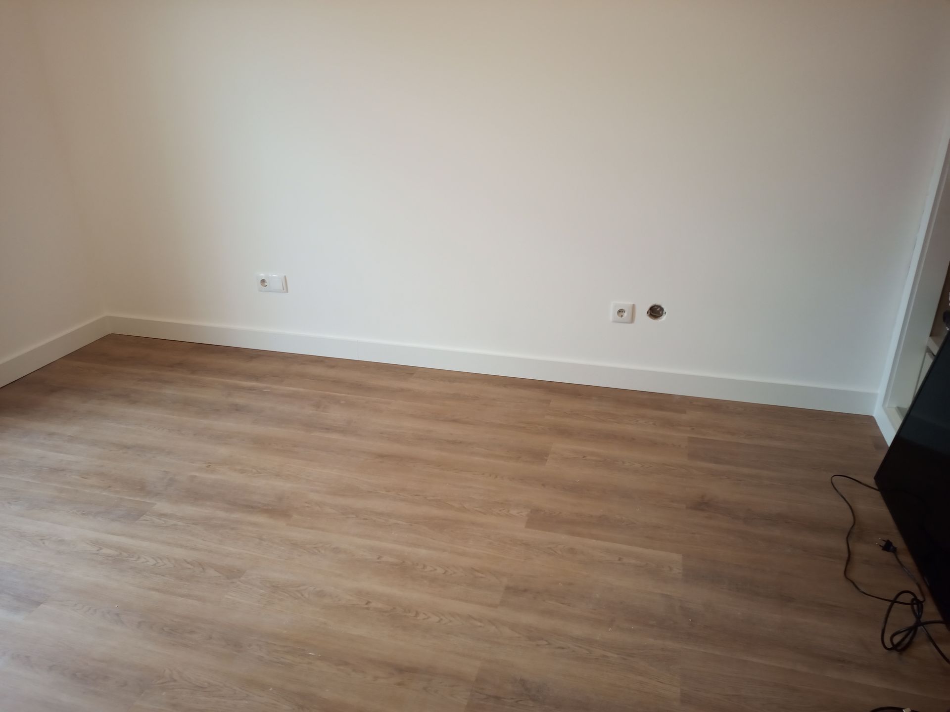 Quarto vazio com piso imitando madeira e paredes brancas. Tomadas elétricas visíveis.