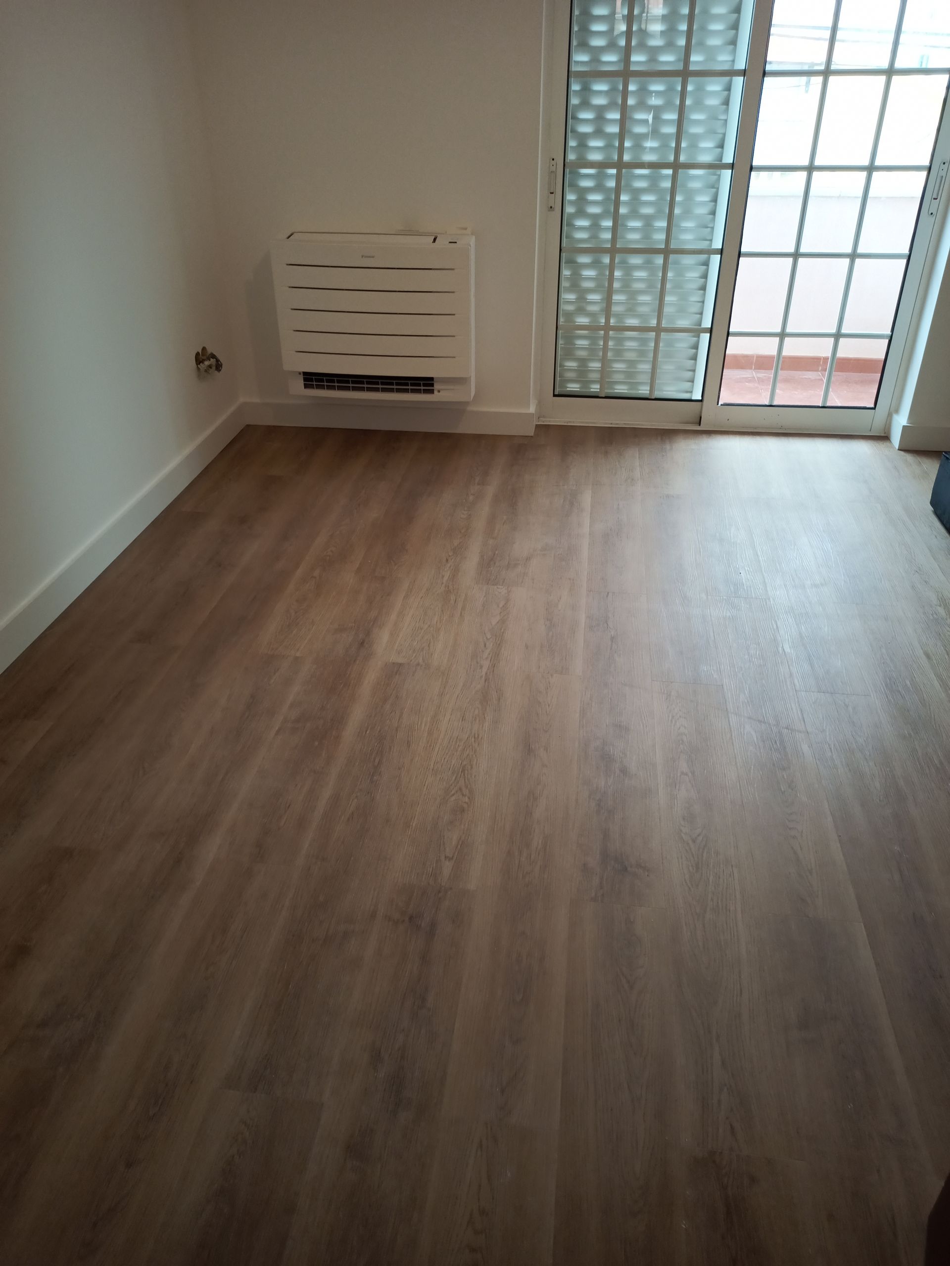 Quarto vazio com piso imitando madeira, paredes brancas, aquecedor e porta de blocos de vidro para uma varanda.