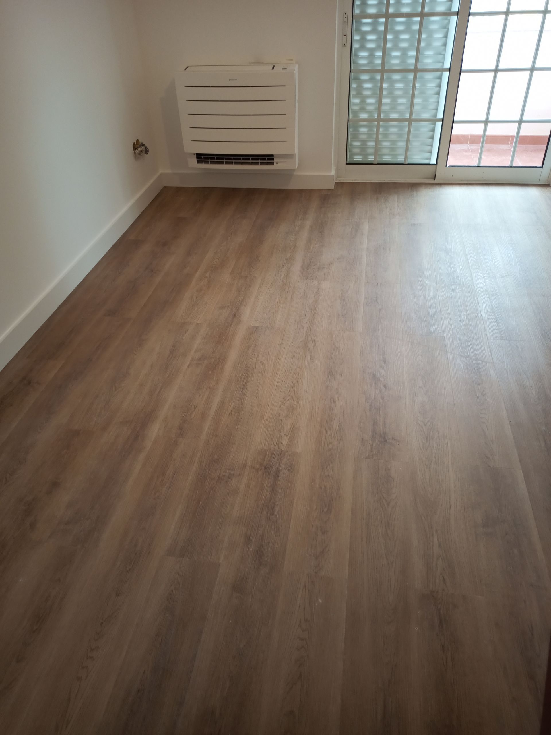 Quarto vazio com piso imitando madeira, acabamentos e paredes brancas, aparelho de ar condicionado e porta de vidro deslizante com grade metálica.