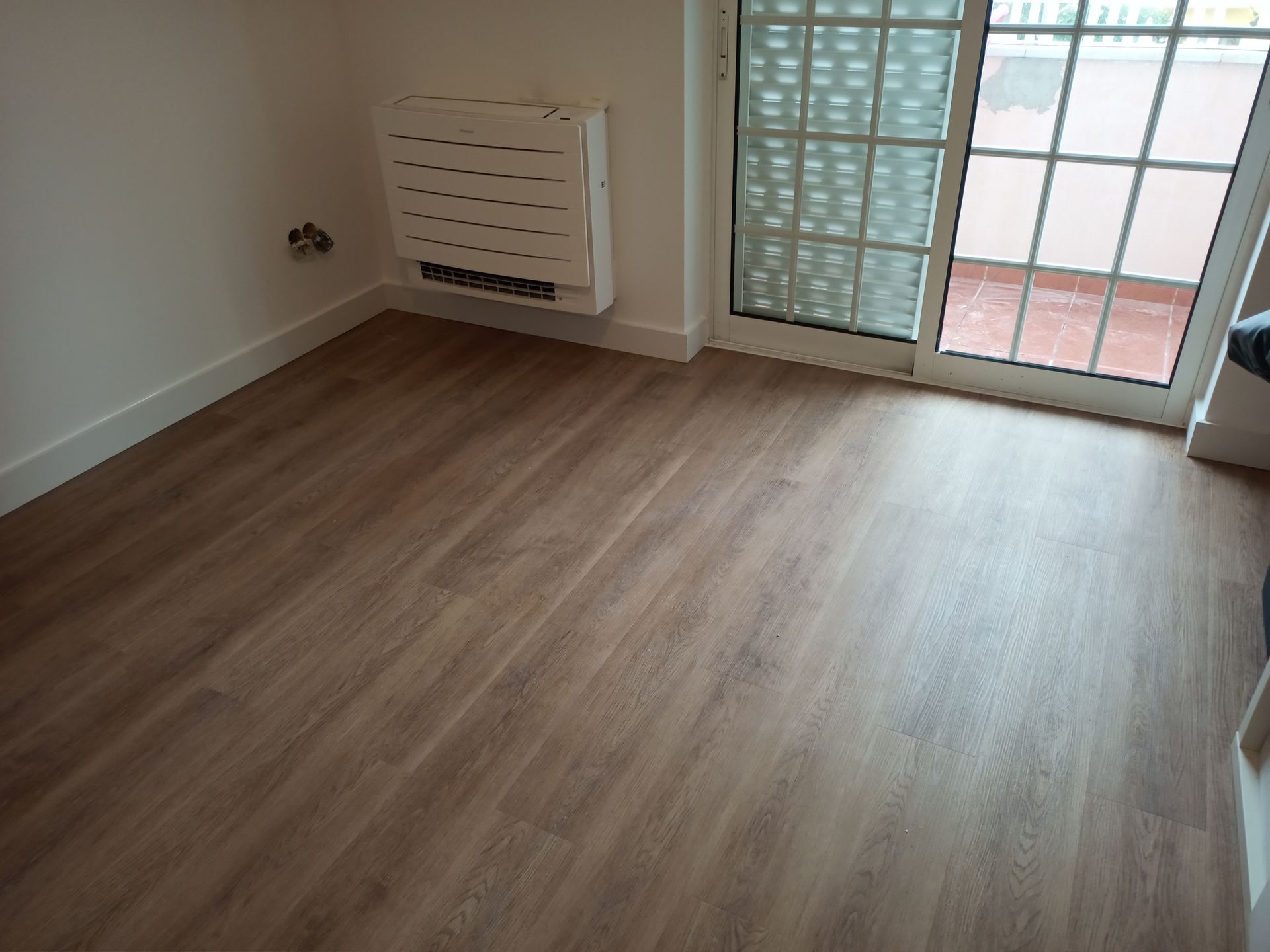 Quarto vazio com piso imitando madeira, paredes brancas e uma porta de vidro deslizante para uma varanda. Há um radiador branco na parede.