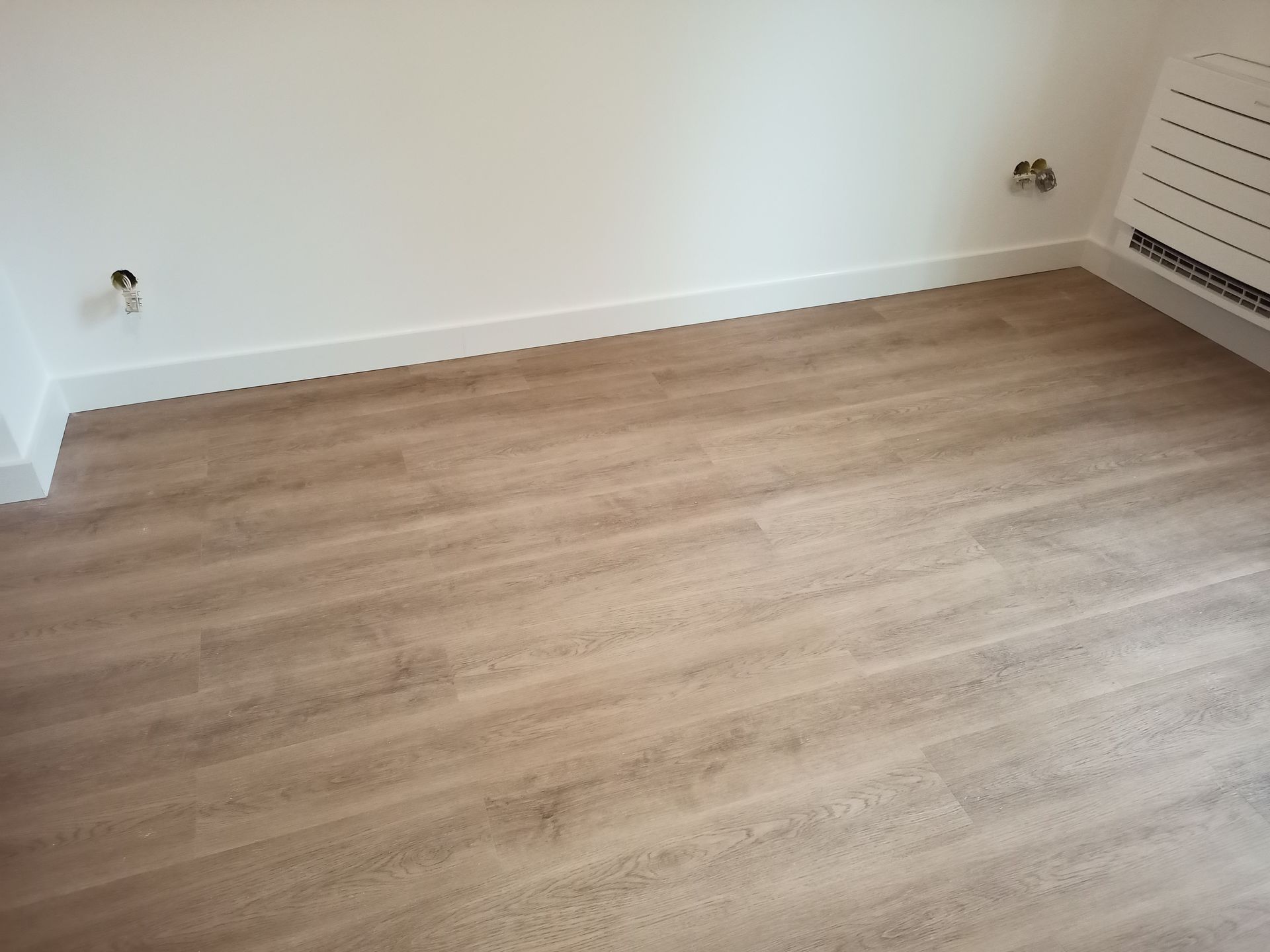 Quarto vazio com piso que imita madeira clara e paredes brancas, rodapé e radiador.