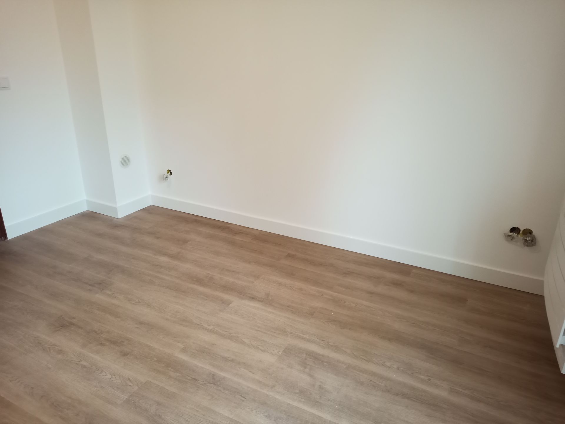 Quarto vazio com piso de madeira marrom-claro, paredes brancas e rodapés.