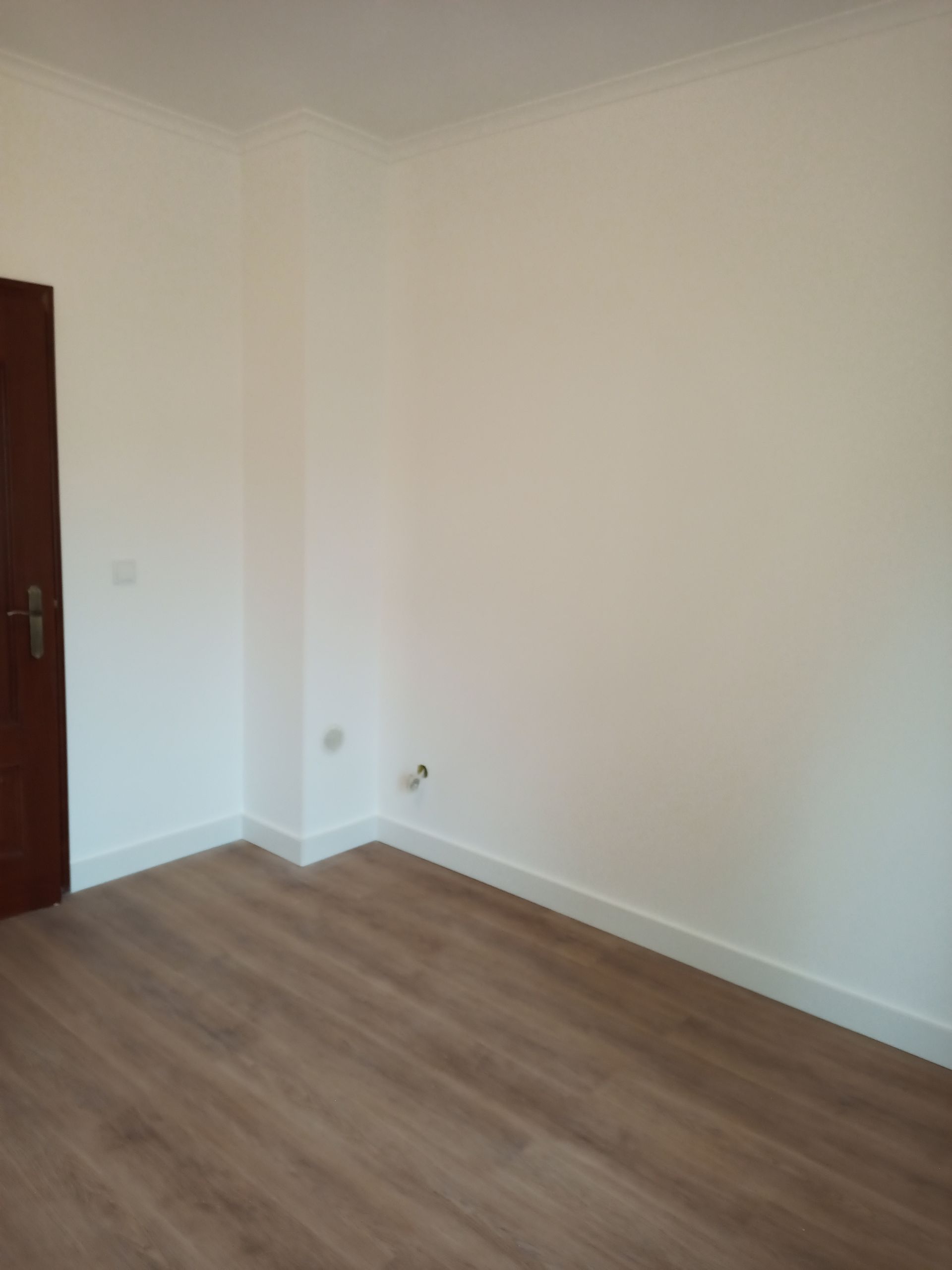 Quarto vazio com piso imitando madeira, paredes brancas, molduras e uma porta marrom.