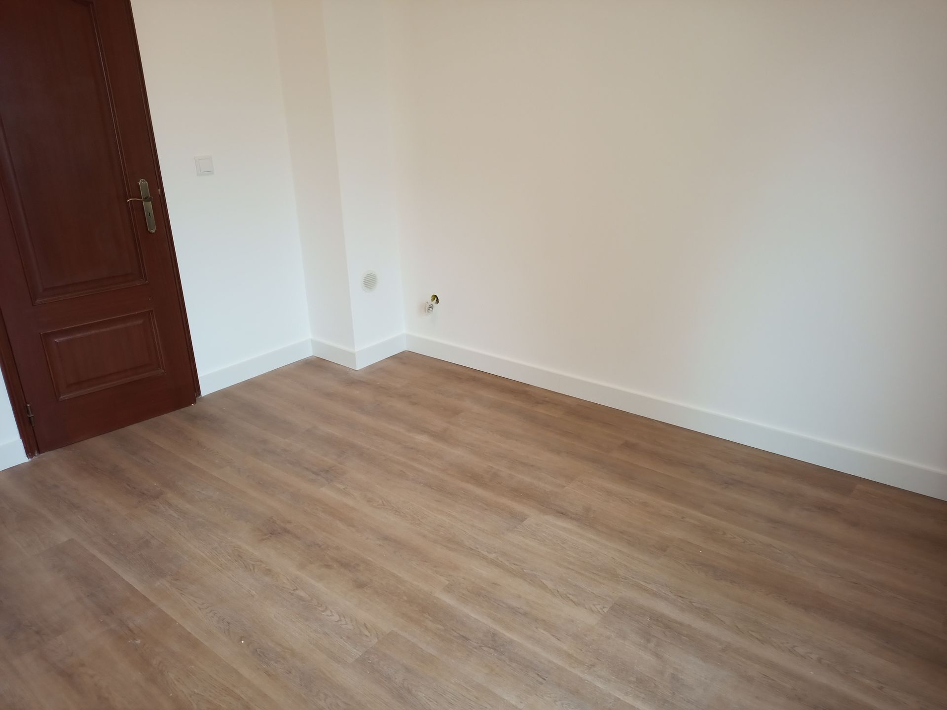 Quarto vazio com piso imitando madeira, paredes brancas e porta marrom.
