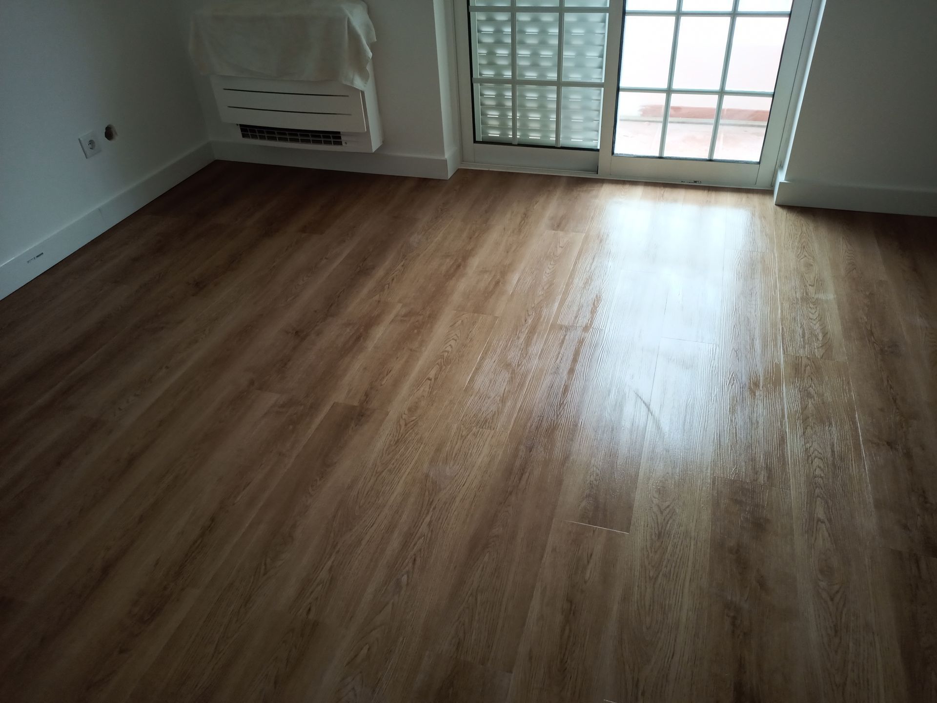 Piso de madeira em um cômodo, com a luz do sol refletindo na superfície. Porta com painéis de vidro à direita.