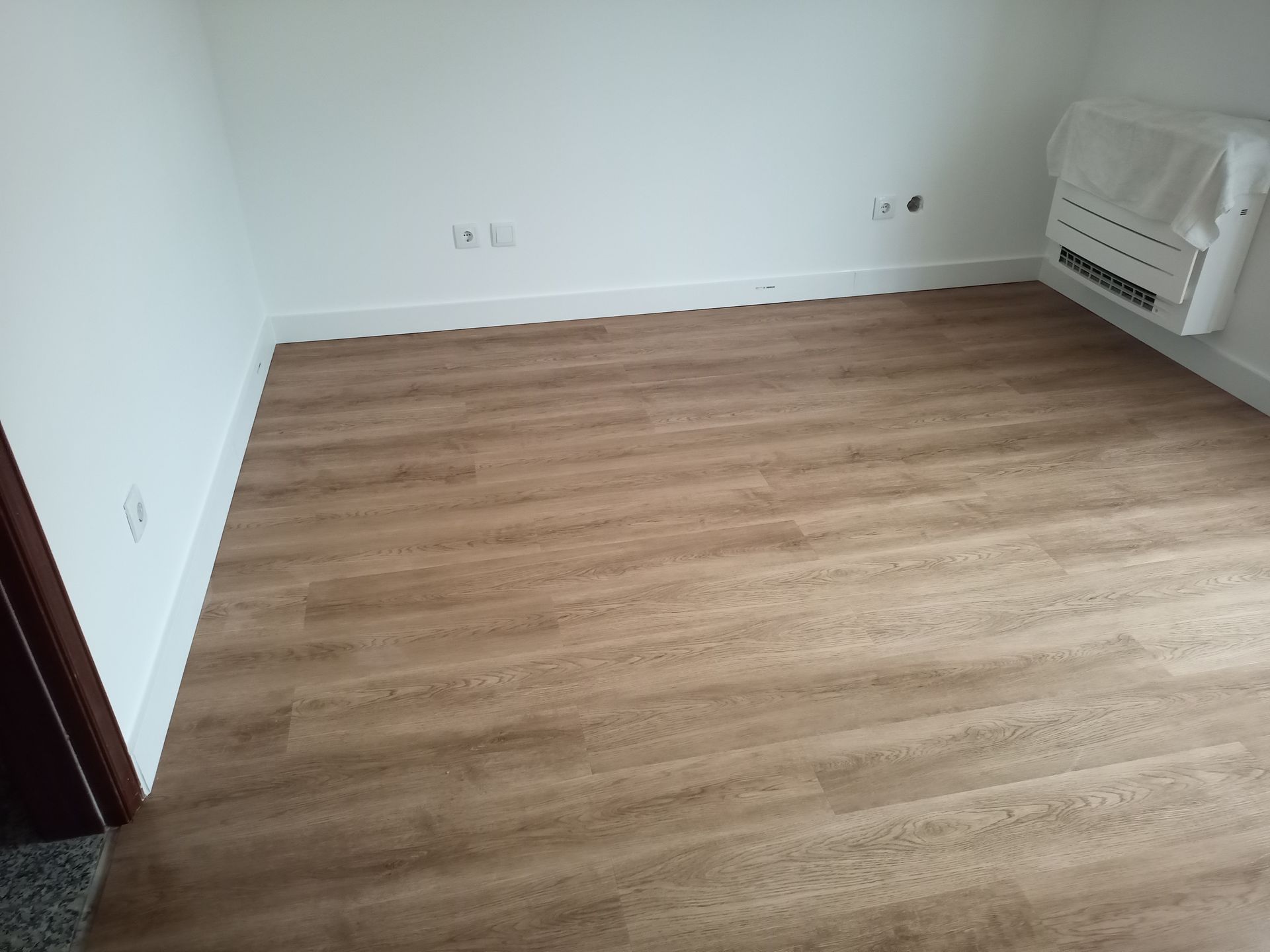 Sala vazia com piso imitando madeira e paredes brancas. Um aquecedor está coberto com plástico.