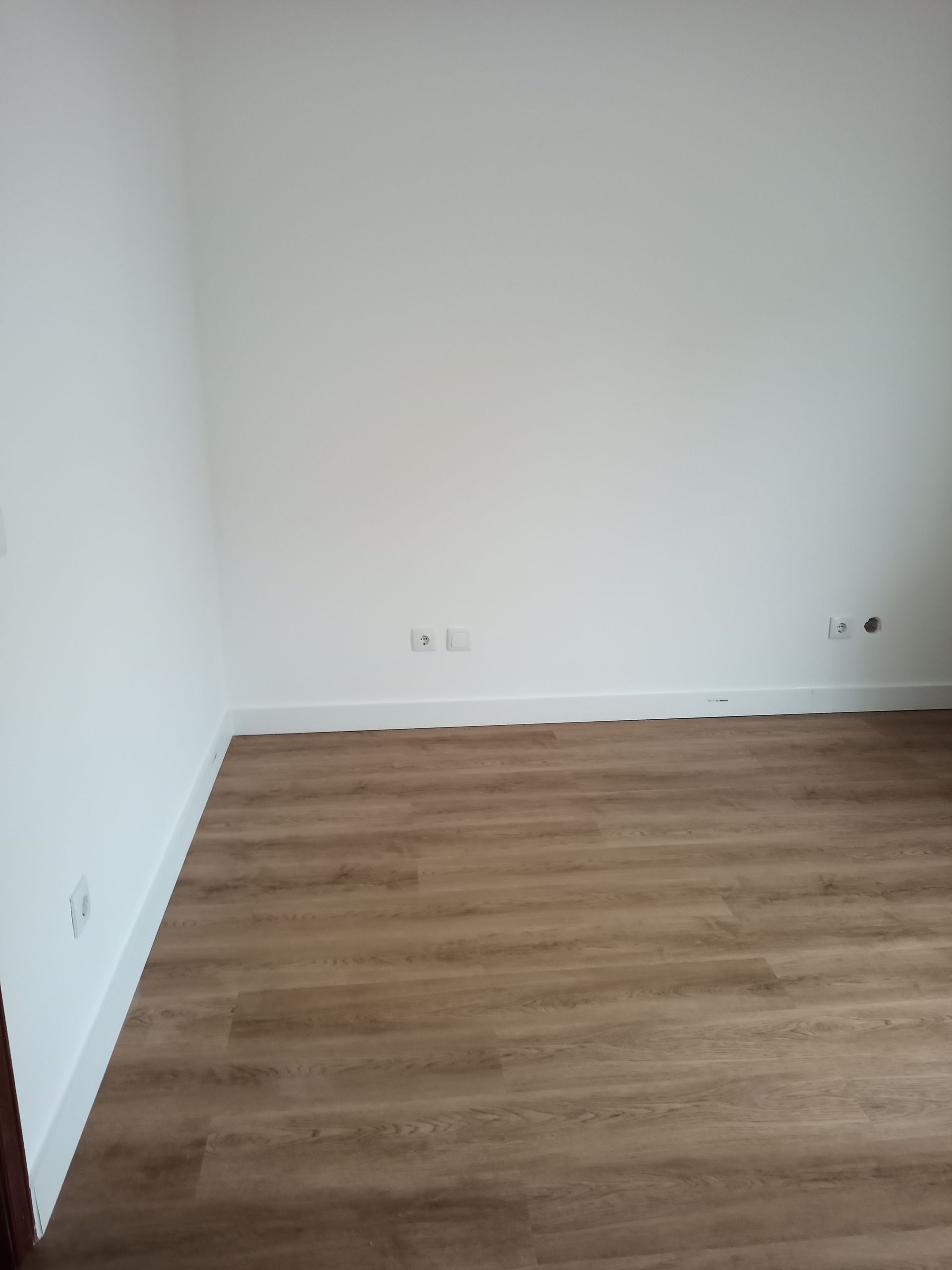 Quarto vazio com paredes brancas, piso imitando madeira marrom e rodapé branco.