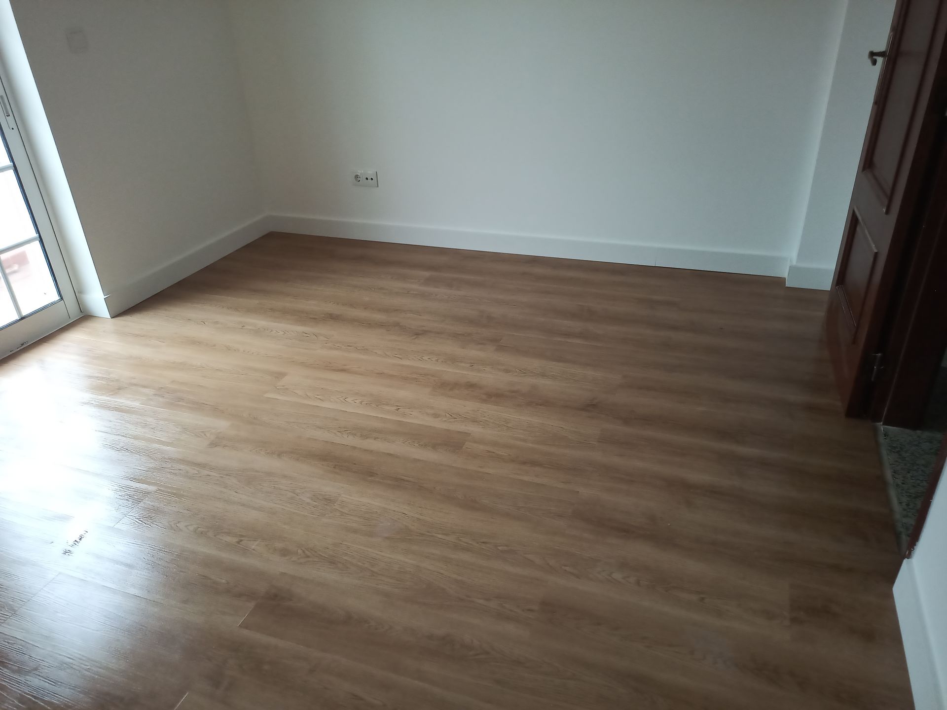 Quarto com piso imitando madeira em tom castanho claro, paredes brancas e uma porta à direita.