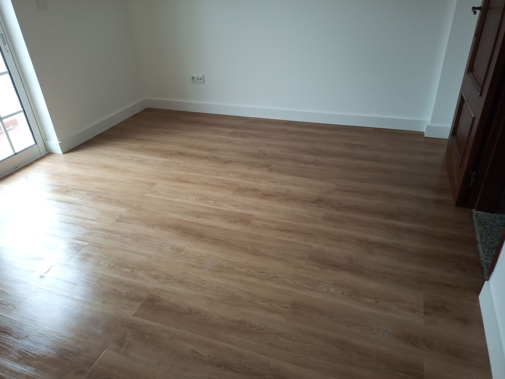 Quarto vazio com piso imitando madeira e paredes brancas. Uma porta e uma janela são visíveis.