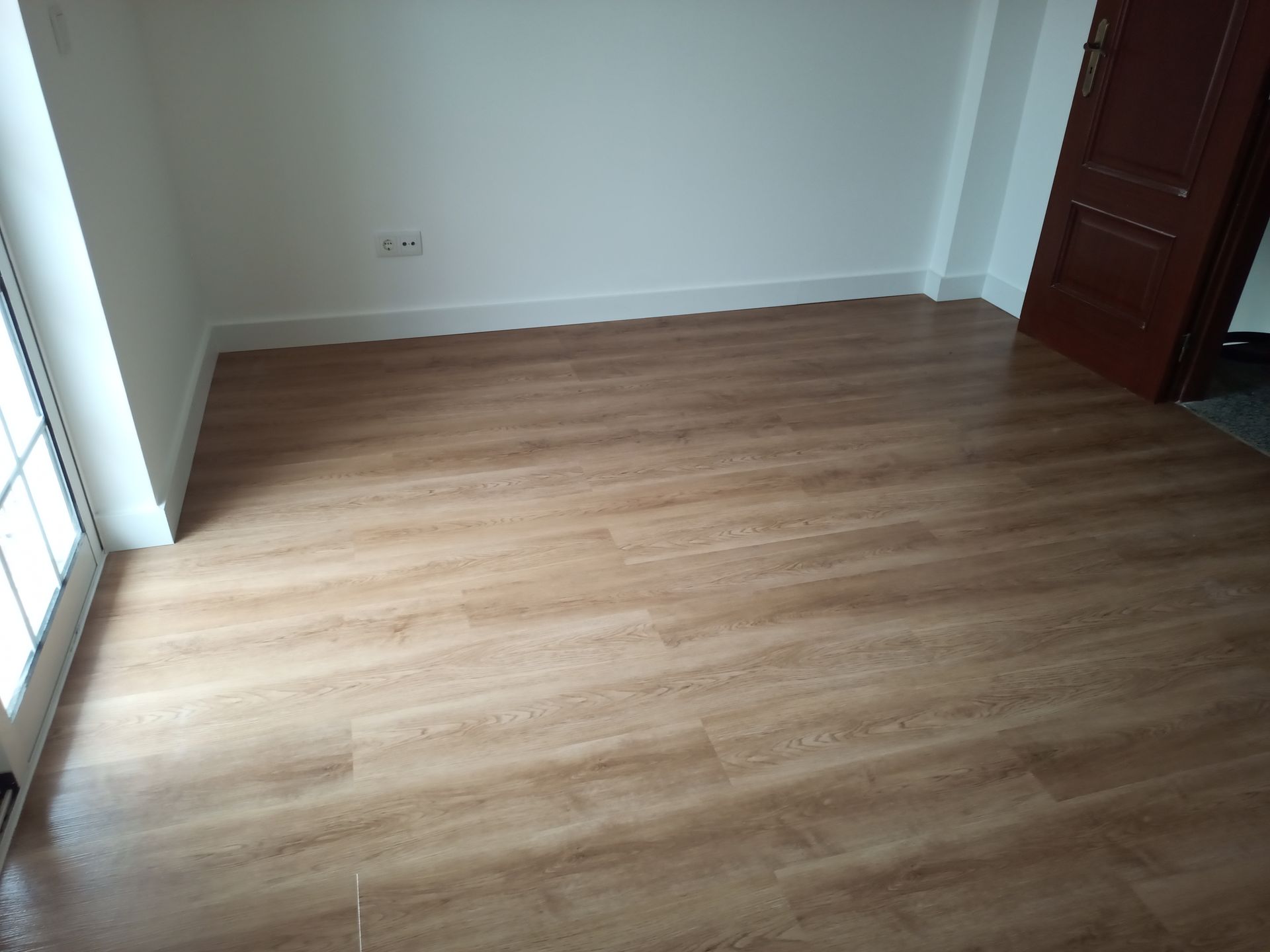 Quarto vazio com piso imitando madeira em tom castanho claro, paredes brancas e uma porta castanha fechada.