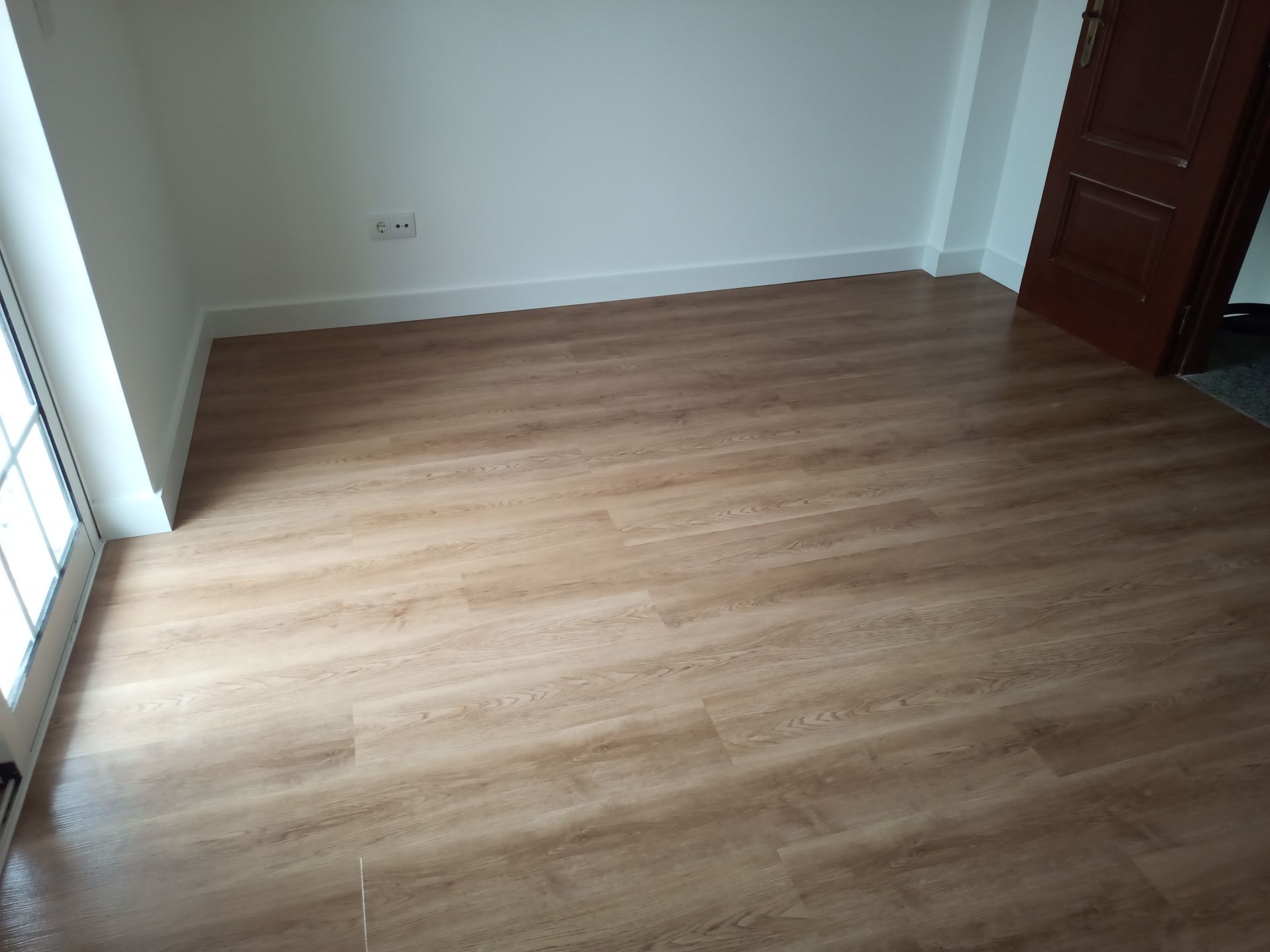 Quarto vazio com piso imitando madeira, paredes brancas e porta marrom.