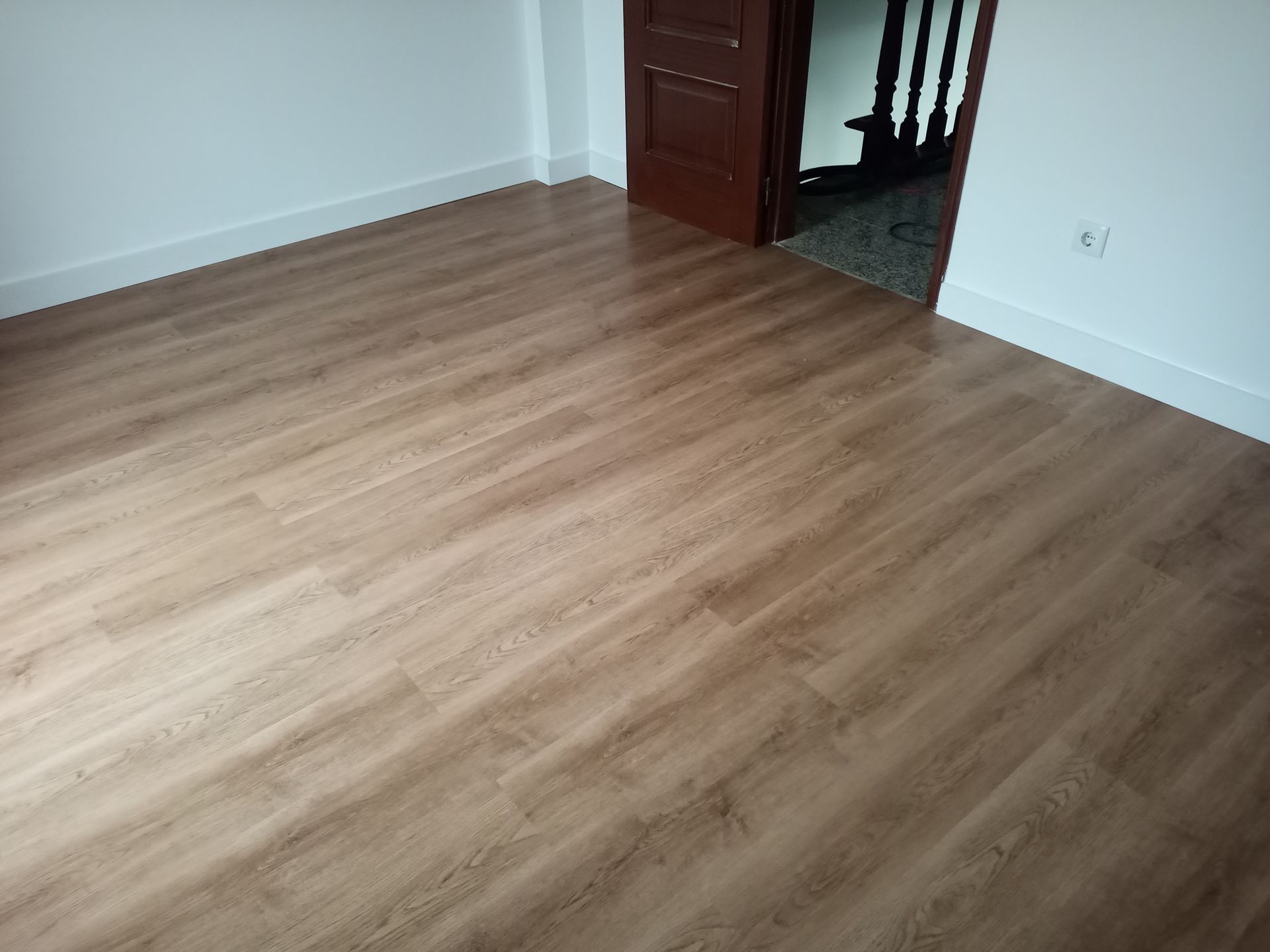 Quarto com piso laminado imitando madeira, paredes brancas e uma porta marrom que dá acesso a uma escada.