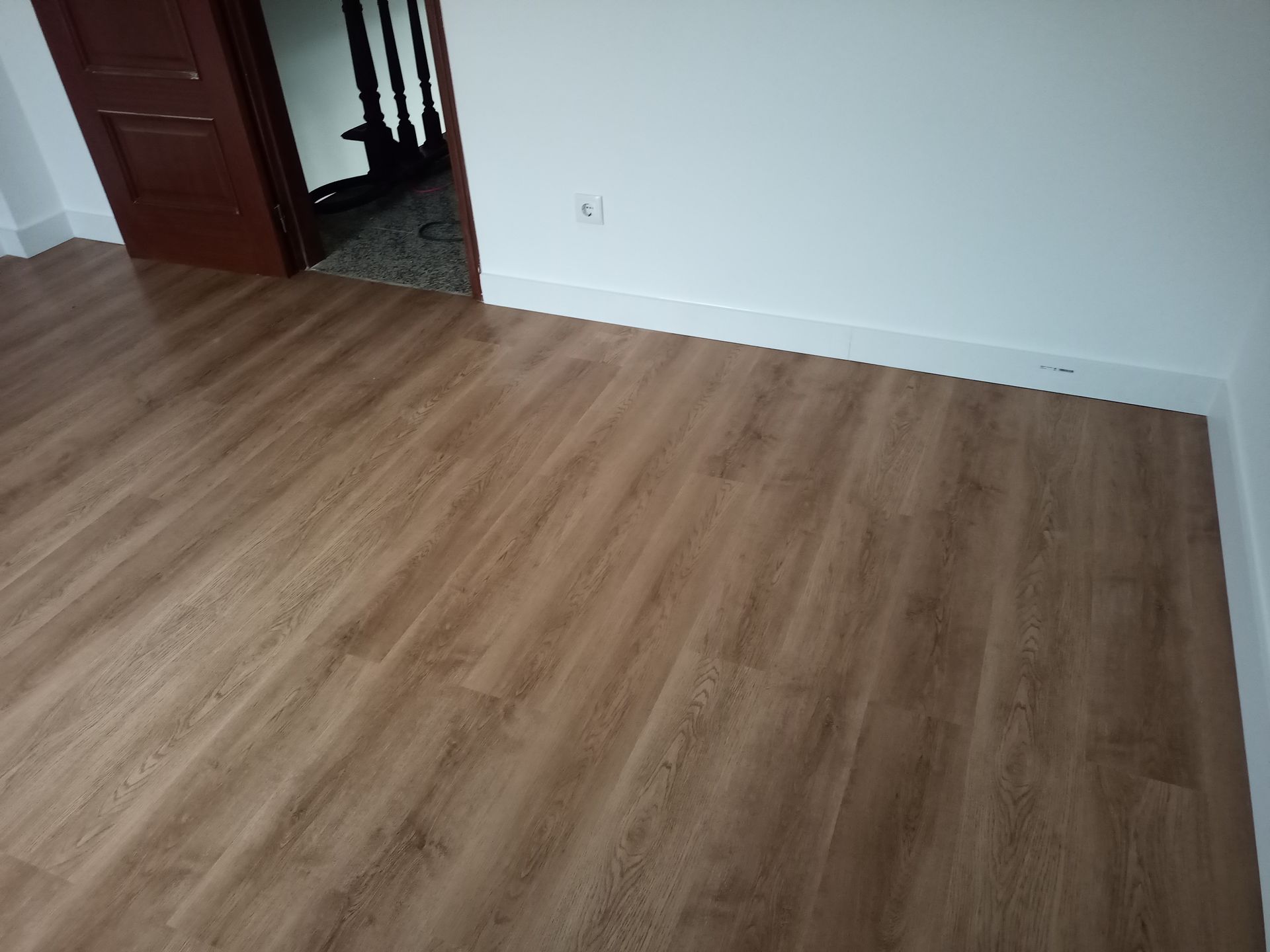 Quarto com piso que imita madeira. Uma porta e a abertura para a escada são visíveis. Paredes e rodapés brancos.