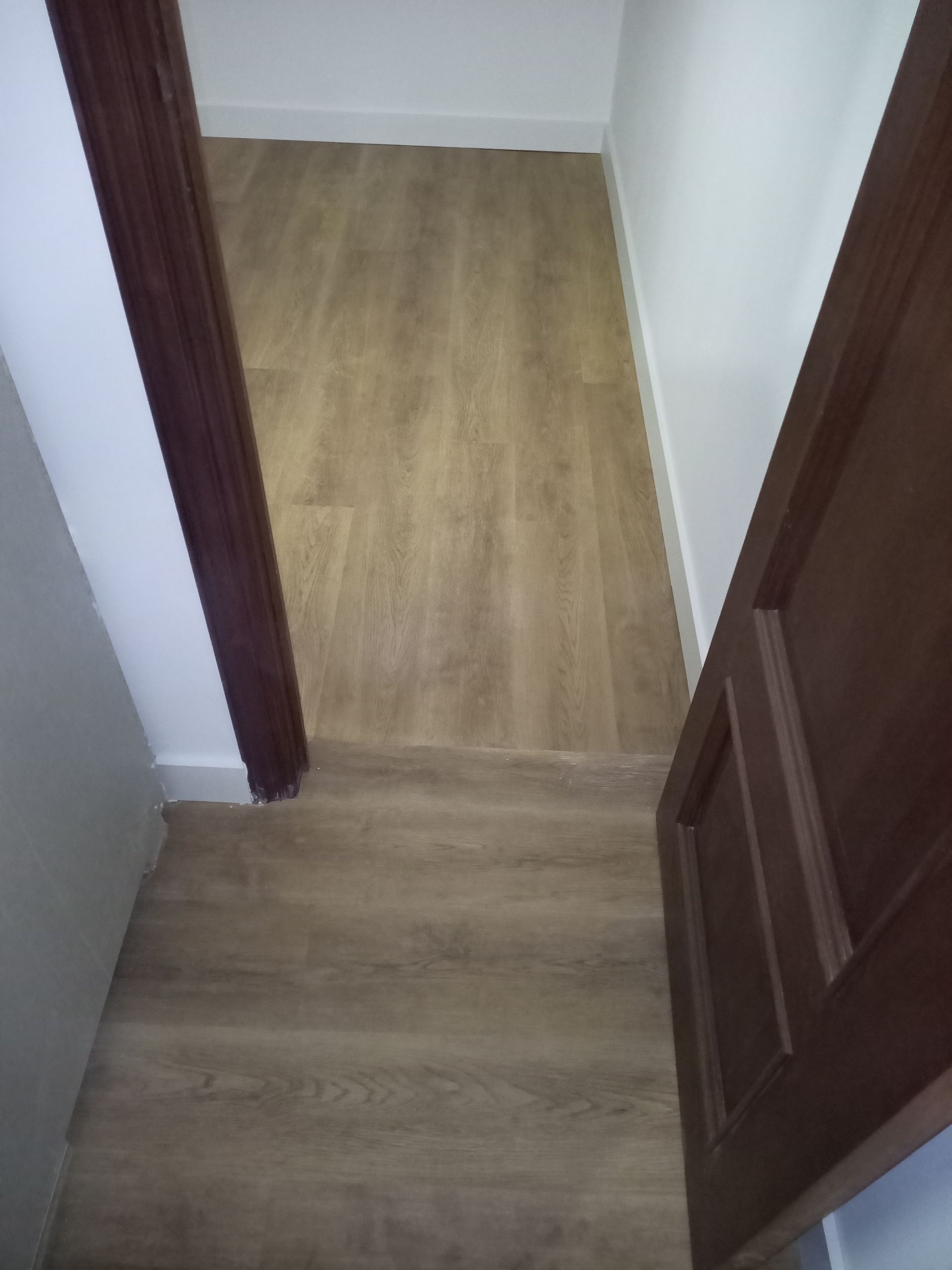 Corredor estreito com piso imitando madeira e uma porta marrom parcialmente aberta à direita. Paredes brancas.