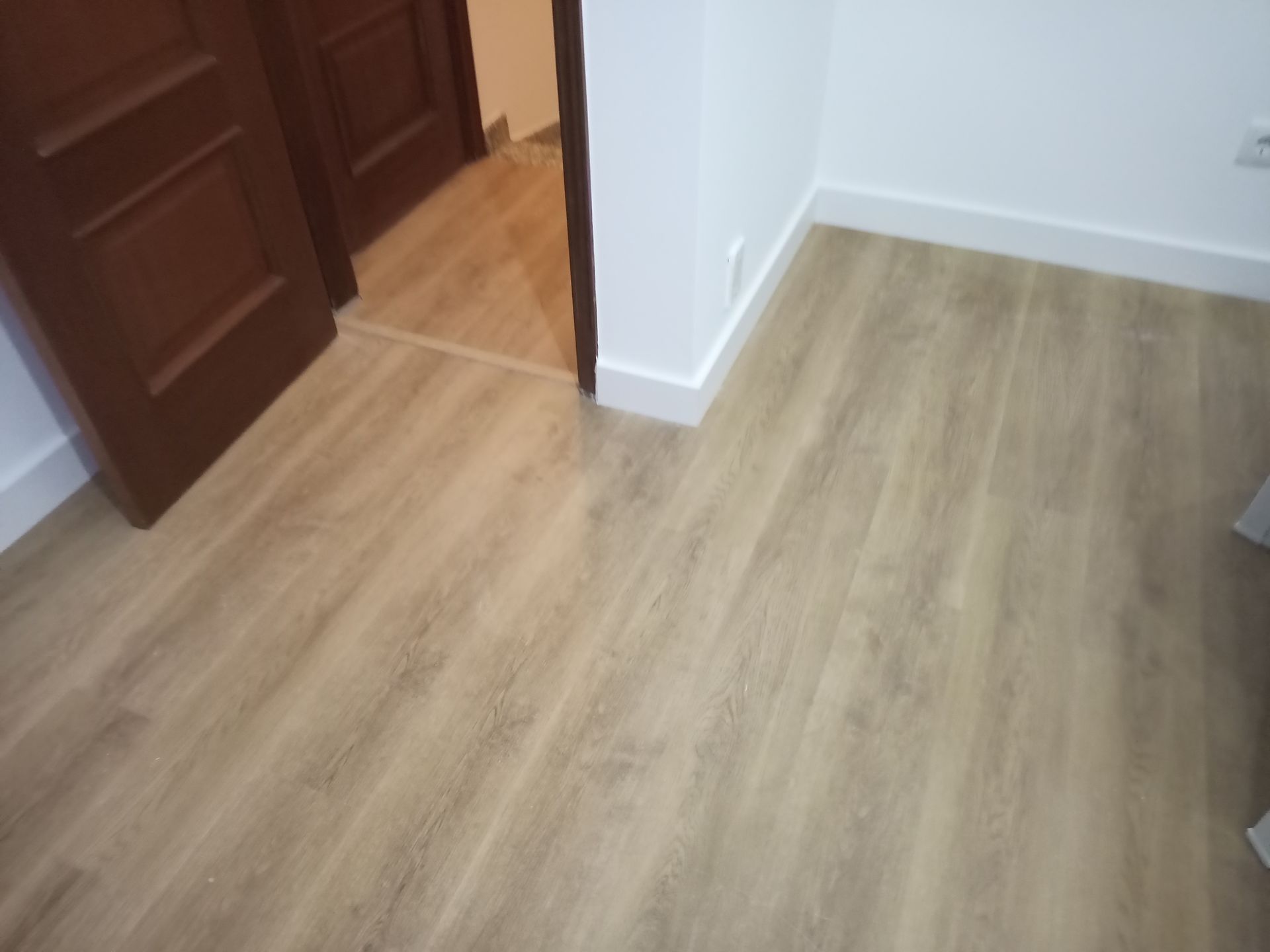 Piso que imita madeira clara e rodapés brancos em um cômodo com porta marrom e vista parcial para um cômodo adjacente.
