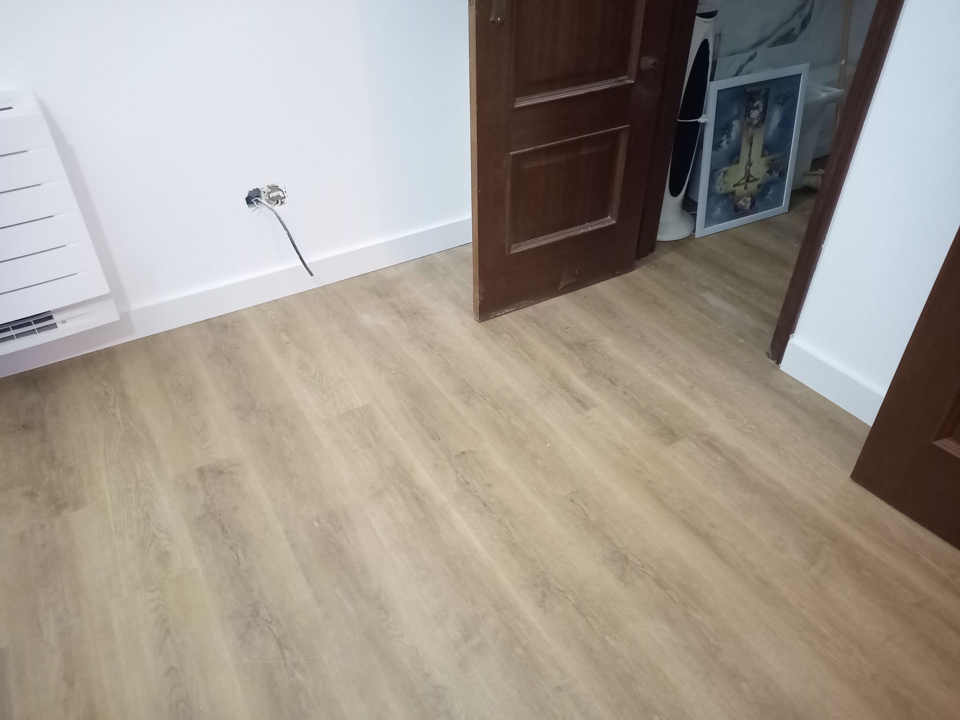 Piso de madeira em um quarto com porta marrom fechada, rodapés brancos e parede branca.